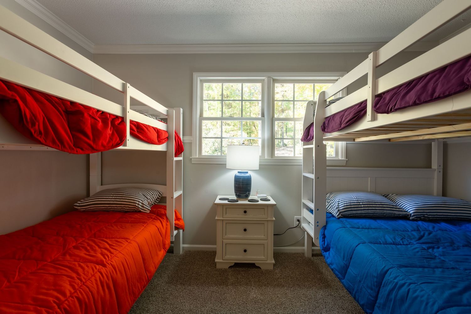A symmetrical bedroom with two bunk beds featuring red and blue bedding and a small nightstand by the window.
