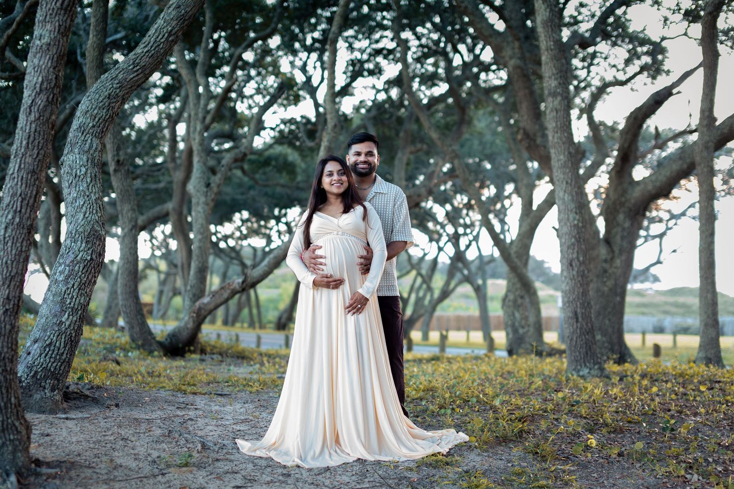 Maternity photo of an expectant couple, the woman in a flowing cream gown, standing under mossy oak trees.