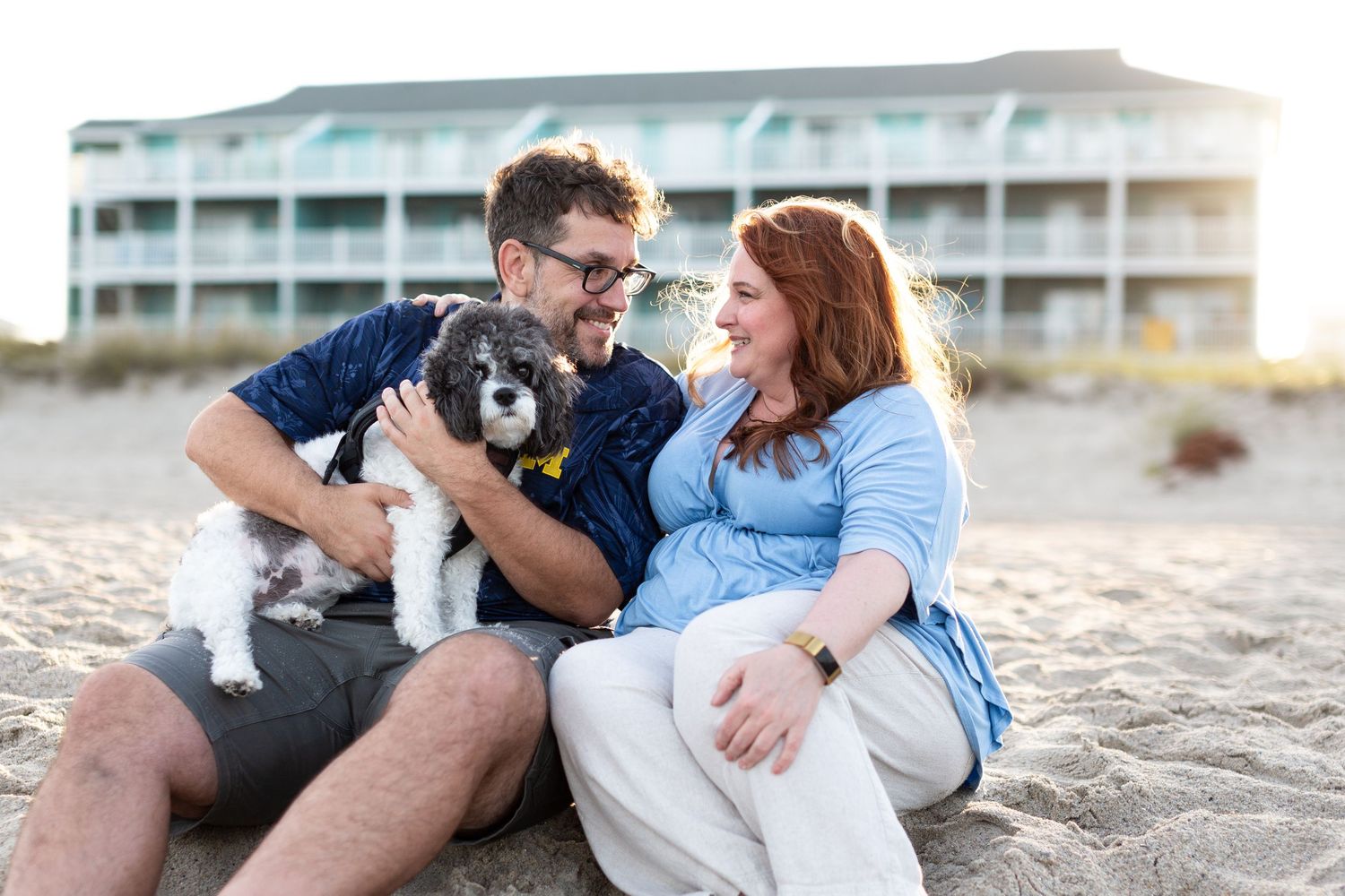Couple sitting on beach with small dog while hotel building visible in background.