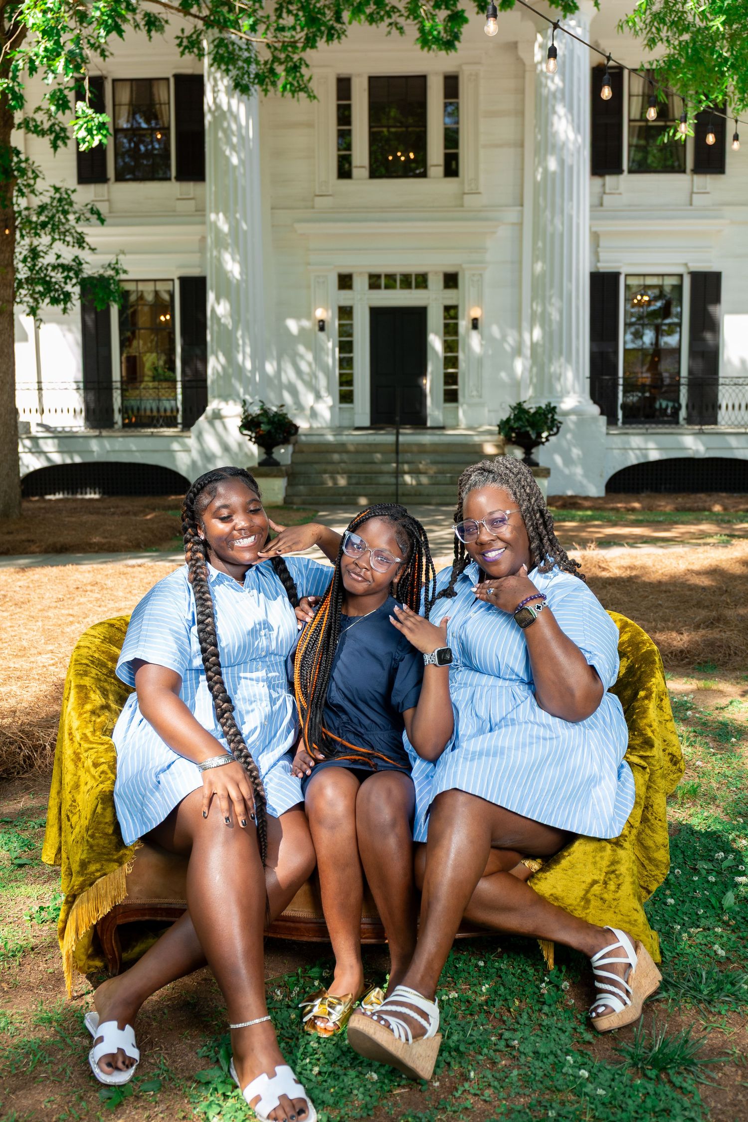 Three young women sitting on a velvet bench outdoors in front of a white colonial mansion, wearing blue dresses.