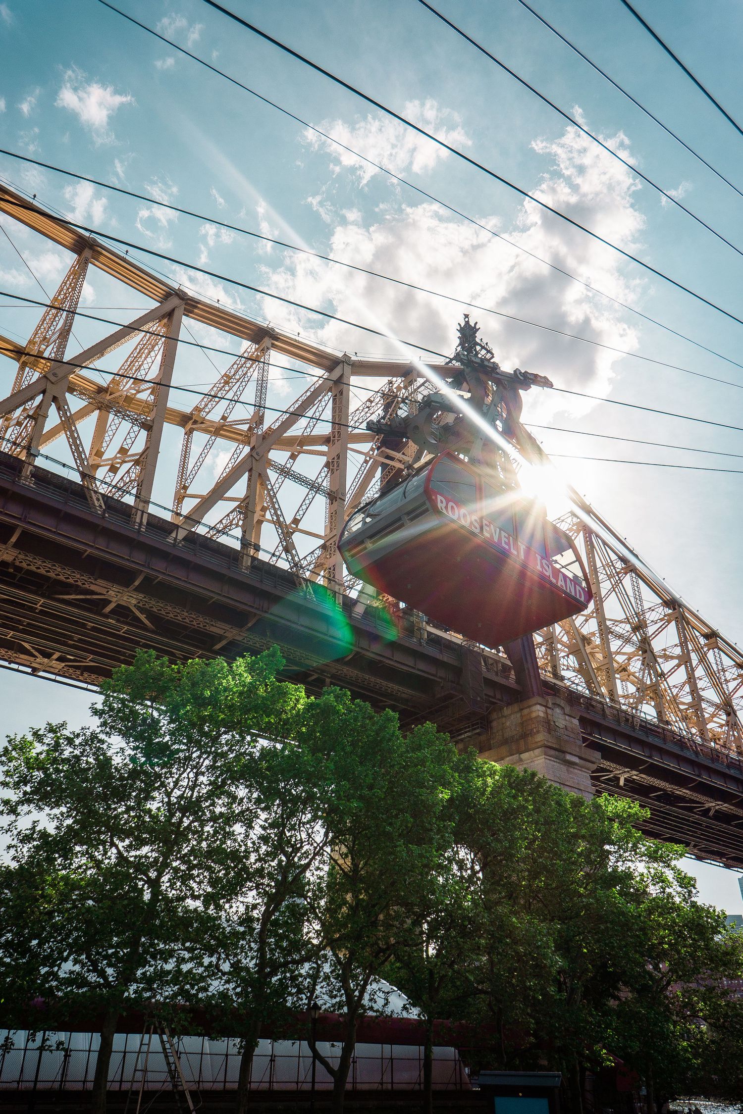 Low angle view of Queensboro Bridge steel structure with sun flare and Roosevelt Island tramway cable car.