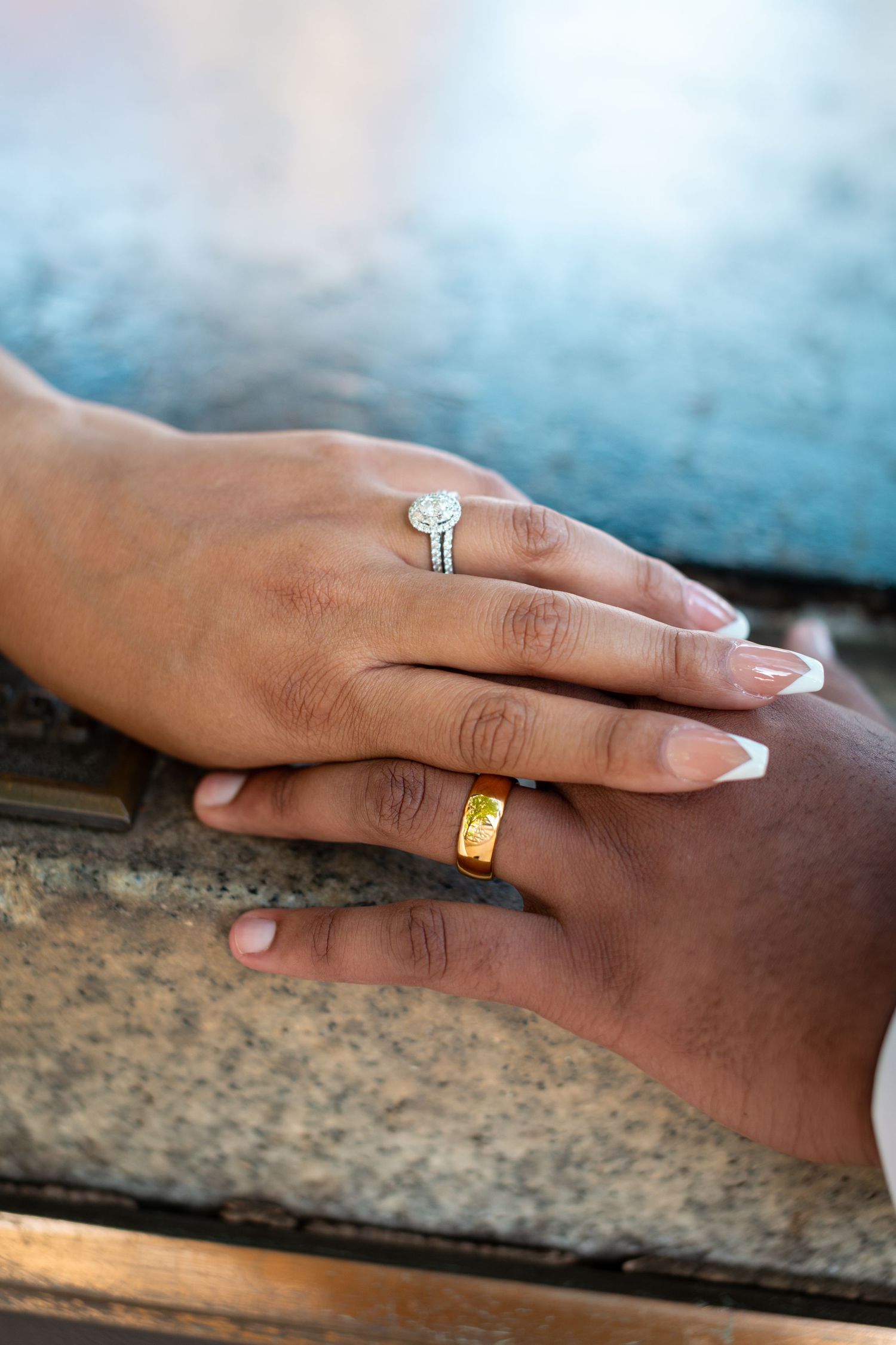 Two hands resting together on granite, wearing matching wedding rings, one silver diamond halo and one gold band.