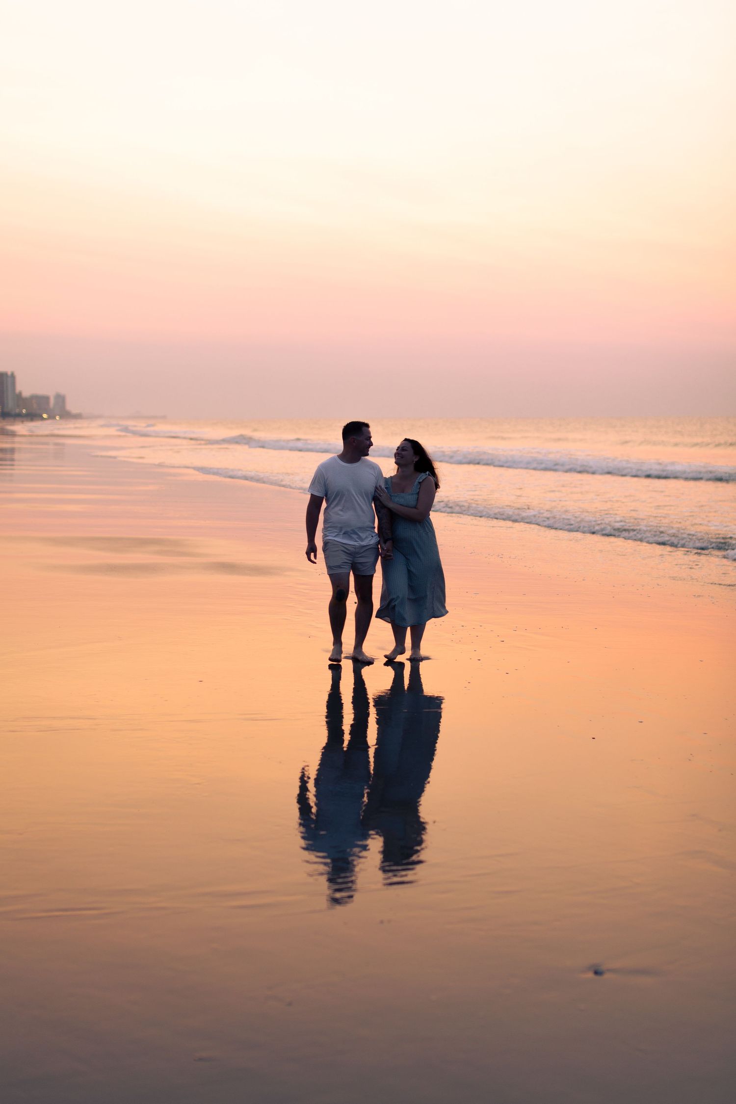 Silhouettes of a couple walking along a beach at sunset with orange and pink sky reflections on wet sand.