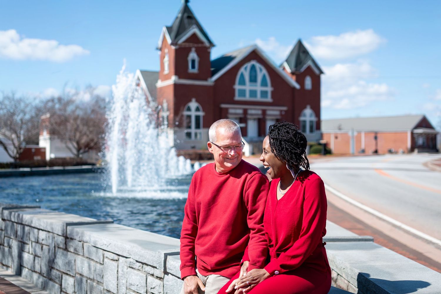 Two people in matching red sweaters sit together by a fountain with a brick church building in the background.