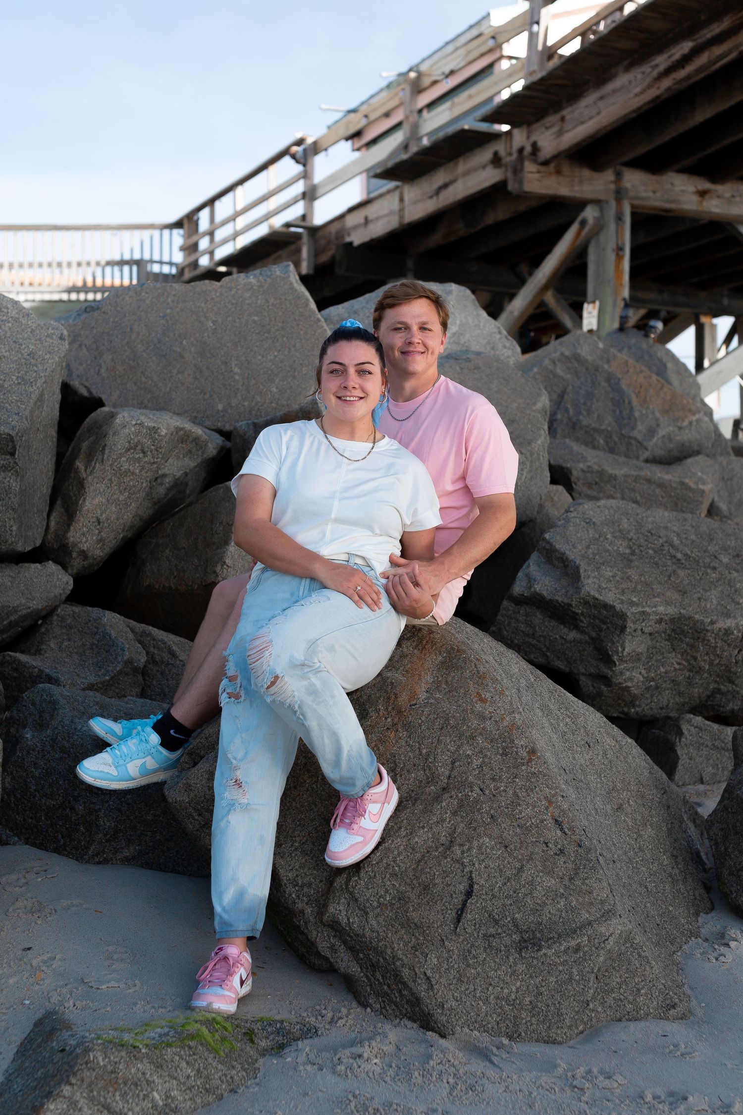 A couple sits together on large rocks at a beach during a casual photo session.