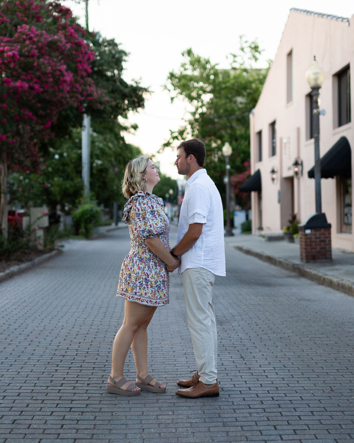 A couple stands on a cobblestone path with blooming bougainvillea flowers overhead on a sunny afternoon.