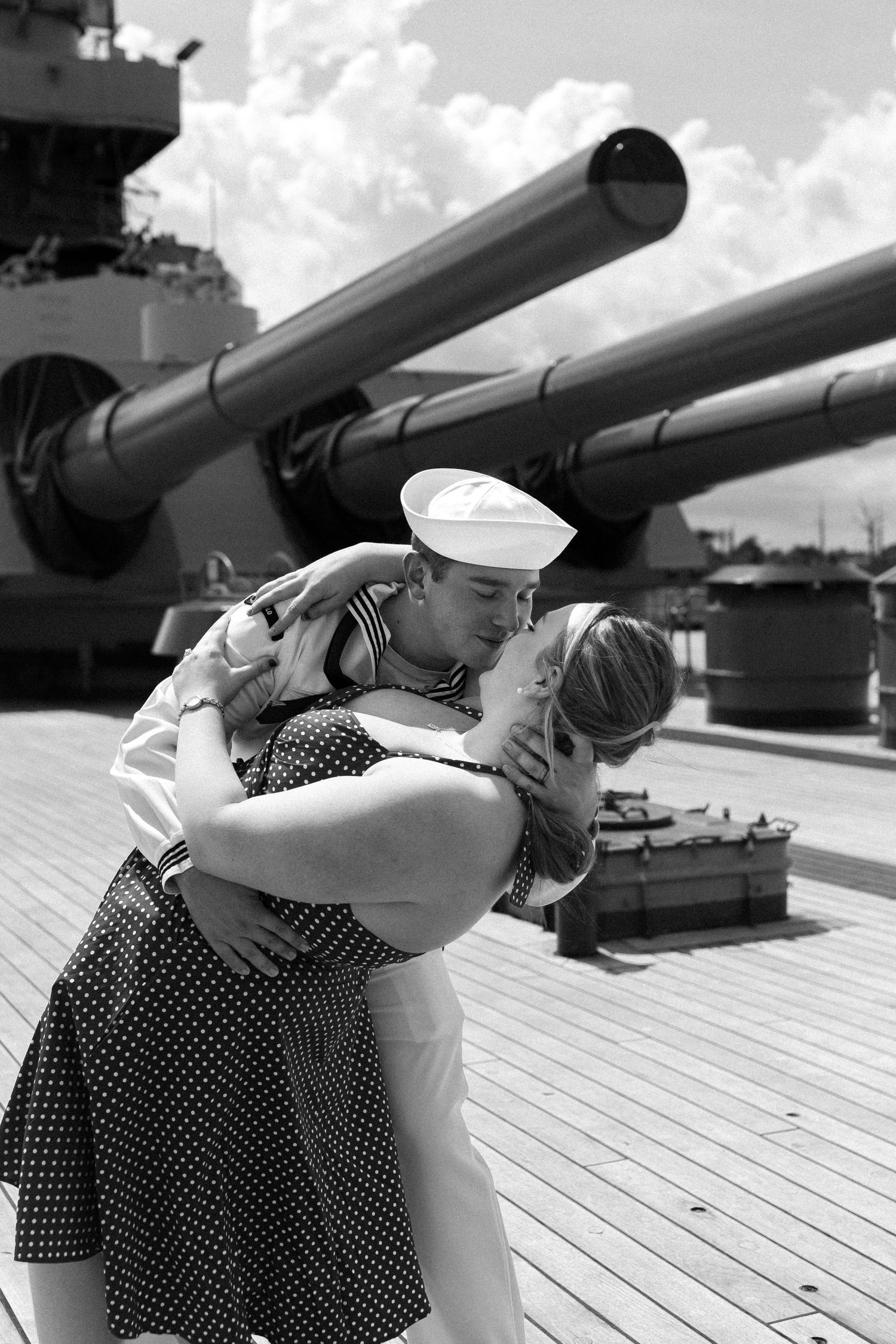 Two sailors in vintage uniforms embrace on a ship deck with naval guns visible in the background.