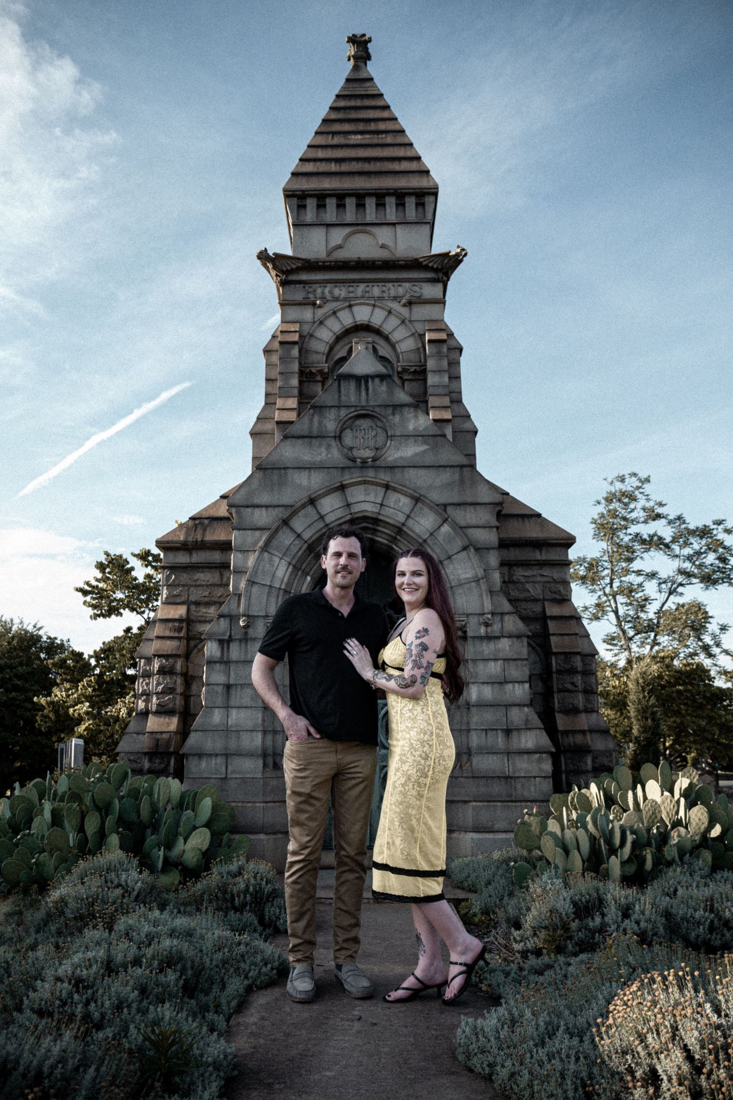 Couple posing in front of a stone monument with cactus garden, man in black shirt and woman in yellow dress.