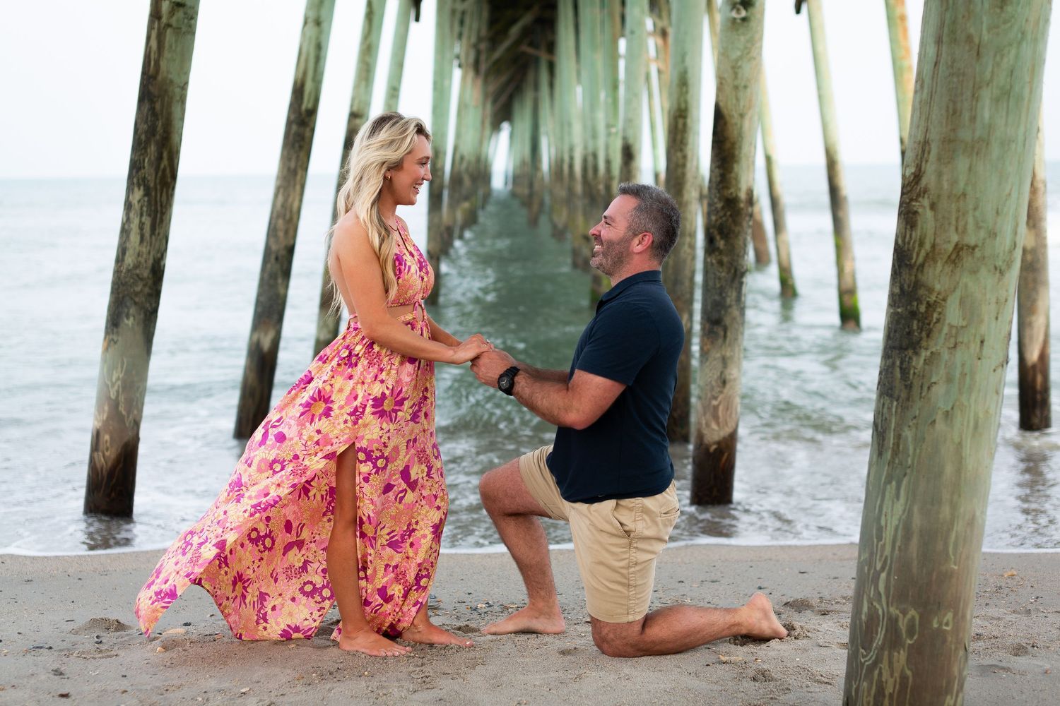 A romantic beach proposal scene under a pier during sunset with waves in the background.
