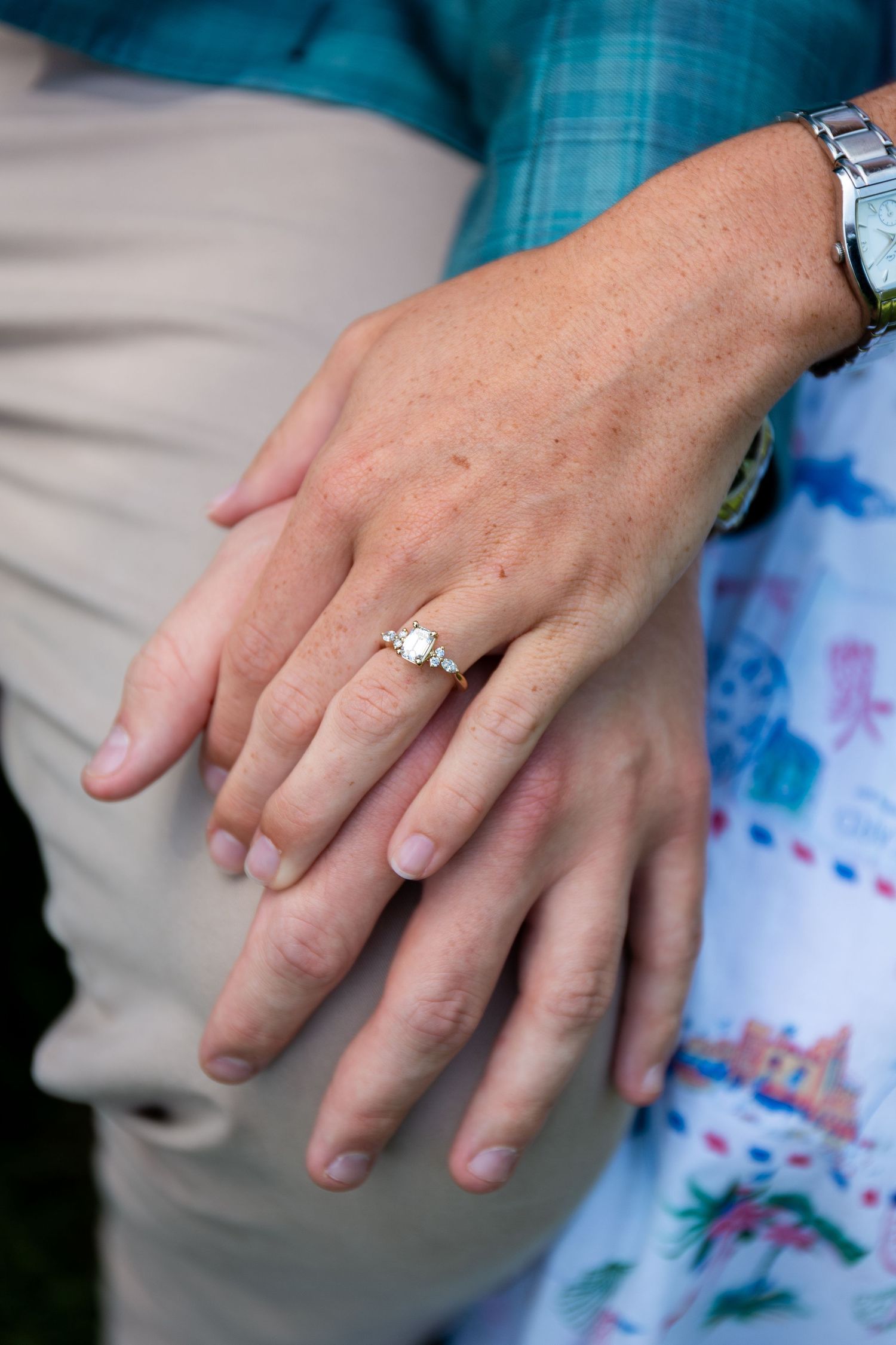 Close-up of hands showing a diamond engagement ring on woman's finger, with man's hand resting beneath hers.