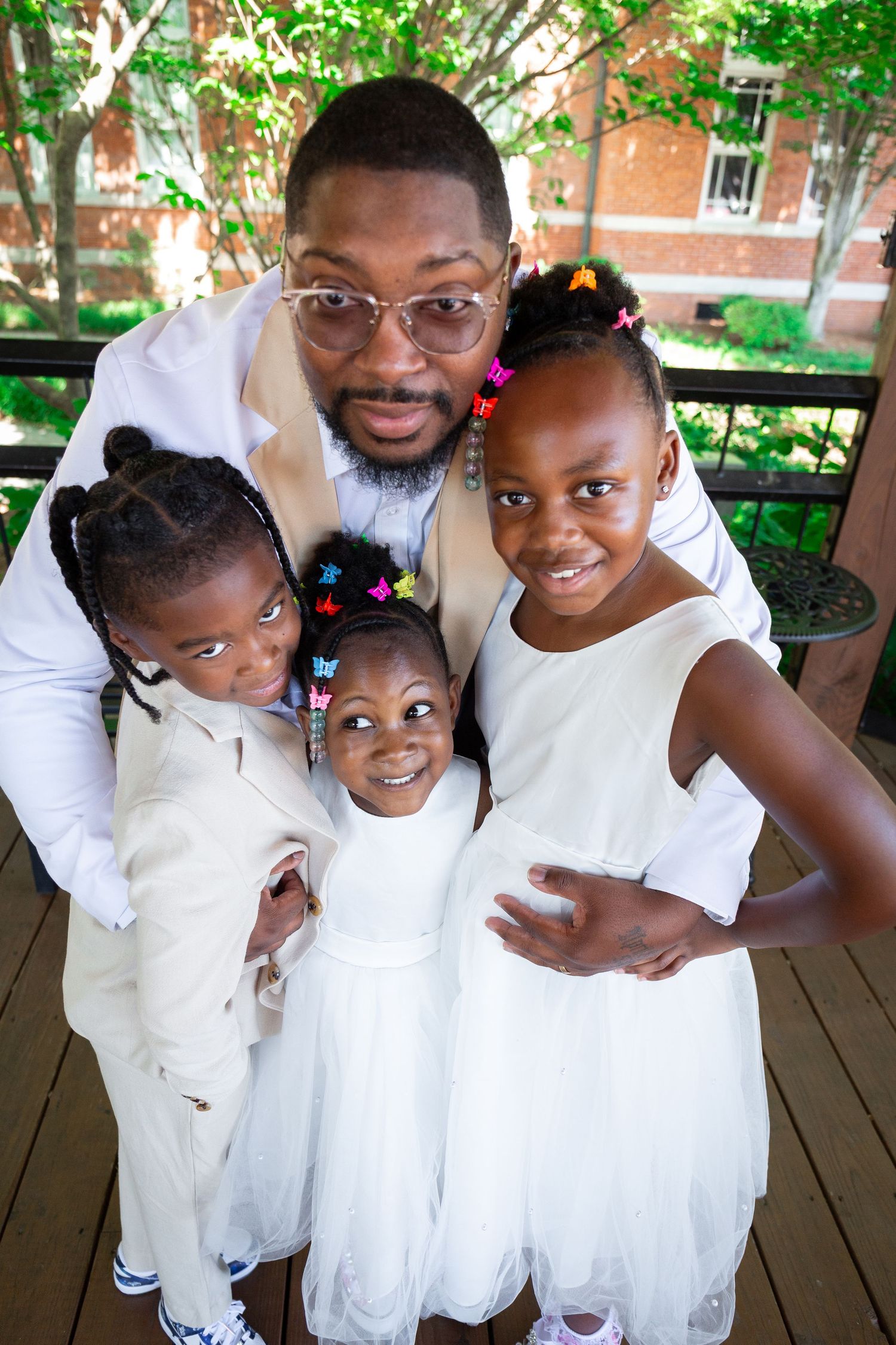 A man in white outfit smiles with three young girls in white dresses on a wooden deck outdoors.