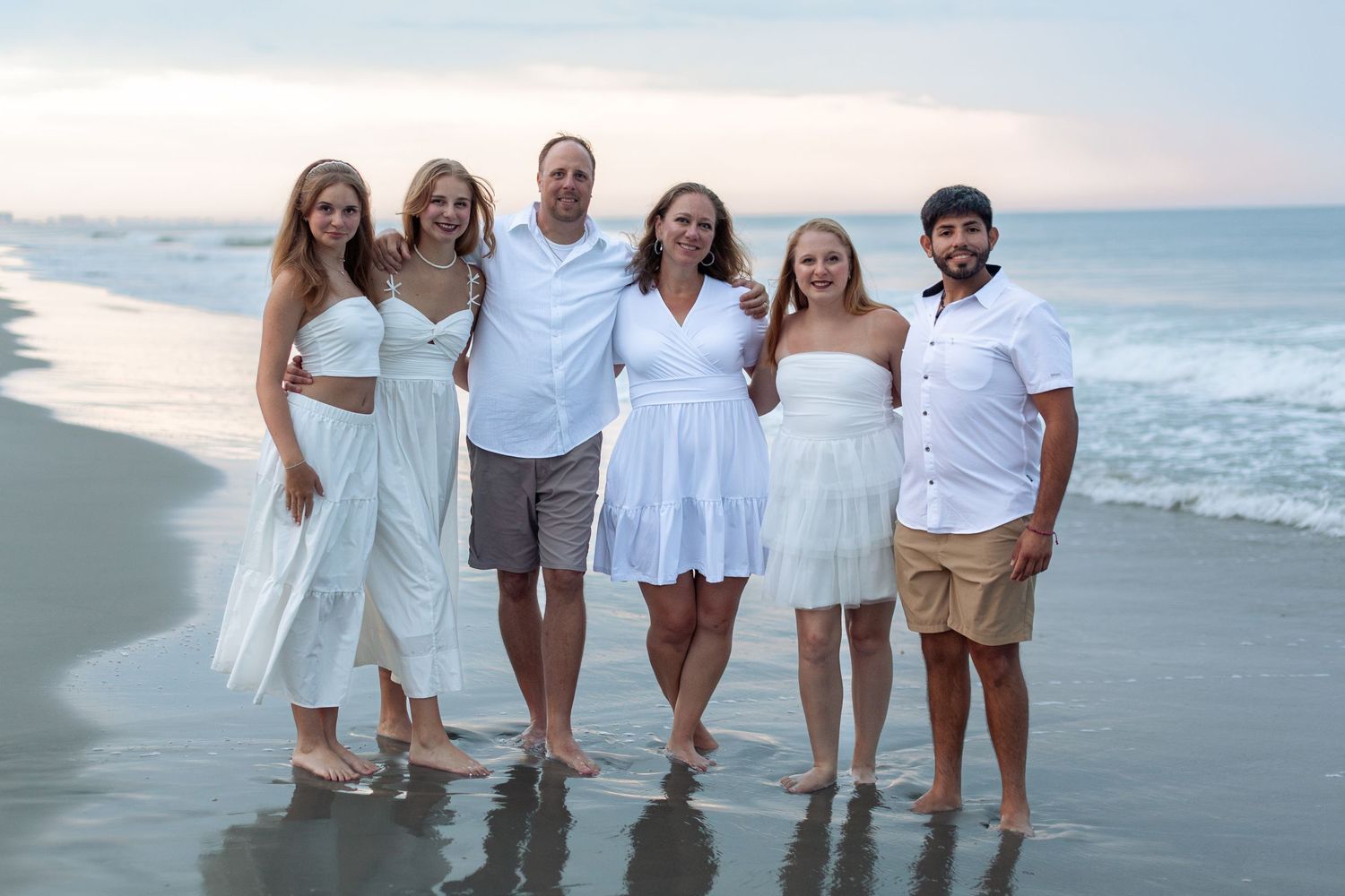 A smiling family of six in white and neutral outfits poses together in shallow water on the beach at sunset.