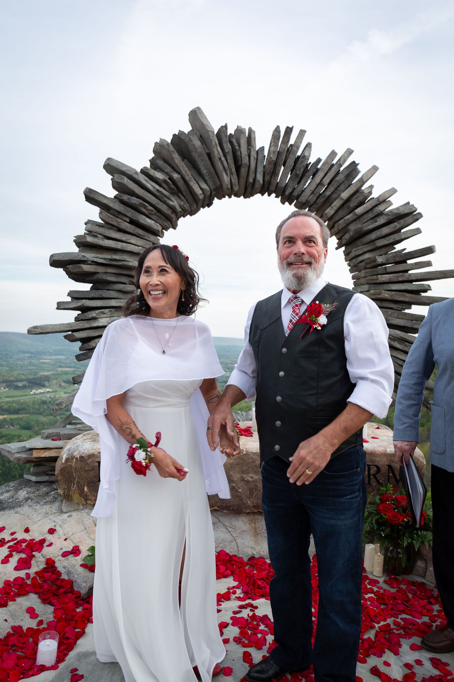 Couple smiling at outdoor wedding ceremony under rustic wooden arch with red rose petals on stone terrace.