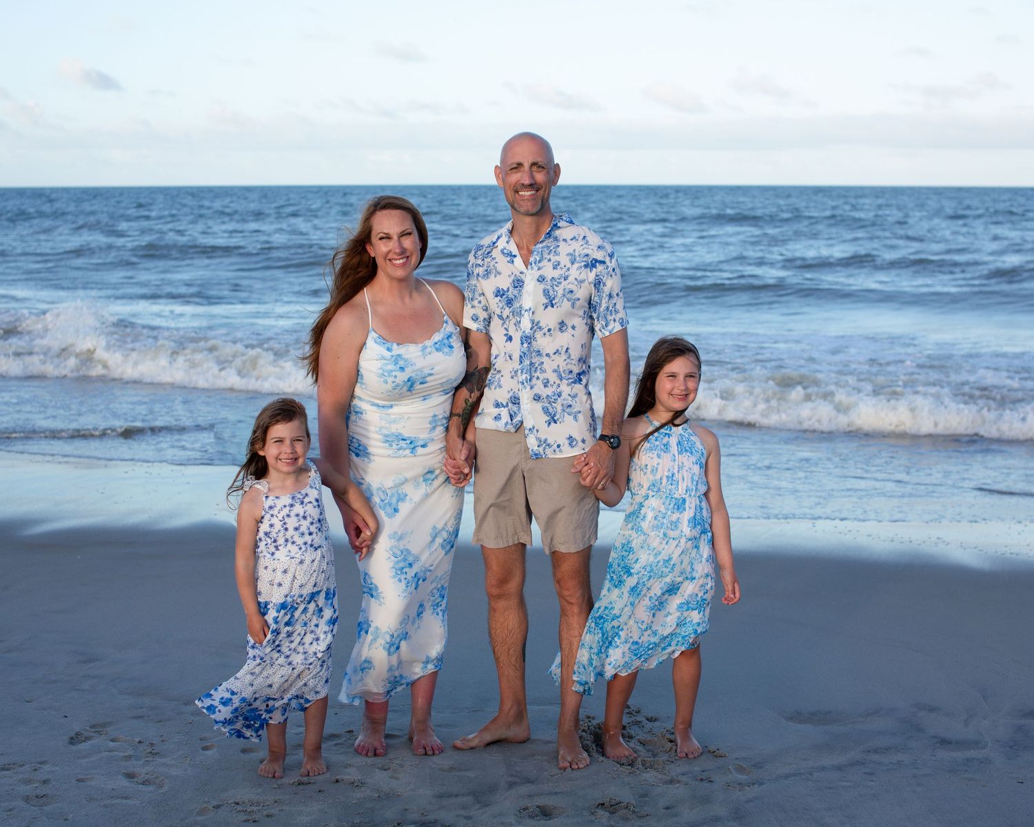 Family in coordinating blue and white outfits posing together on beach at sunset.