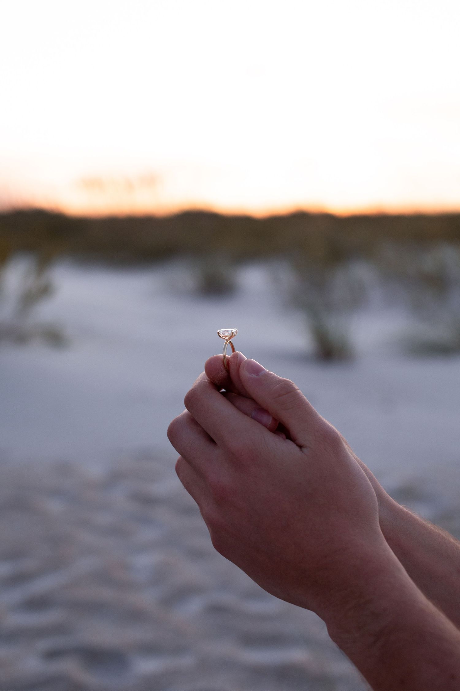 Hands hold up an engagement ring against a sunset beach backdrop.