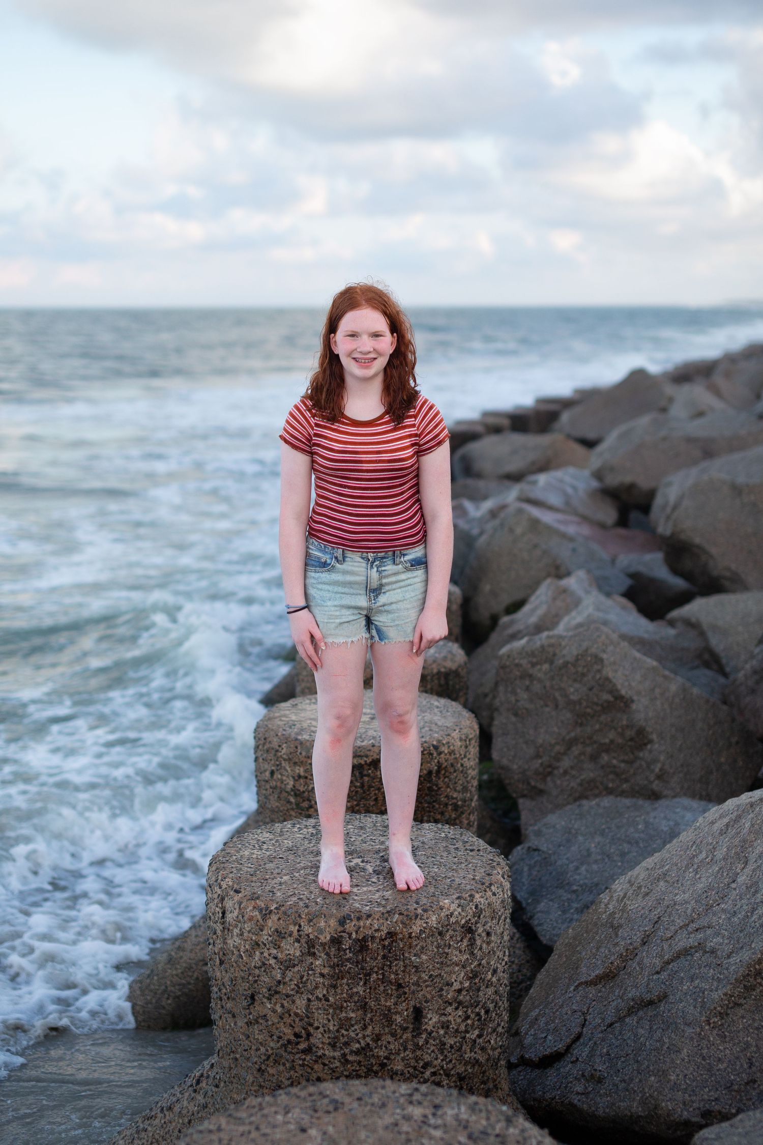 Young person in striped shirt and shorts standing on rocky coastal breakwater at sunset.
