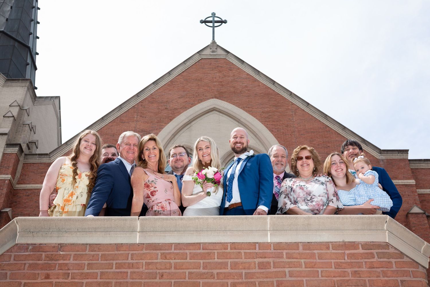 Wedding party posing on church steps in front of brick building with cross, bride in white gown holding bouquet.
