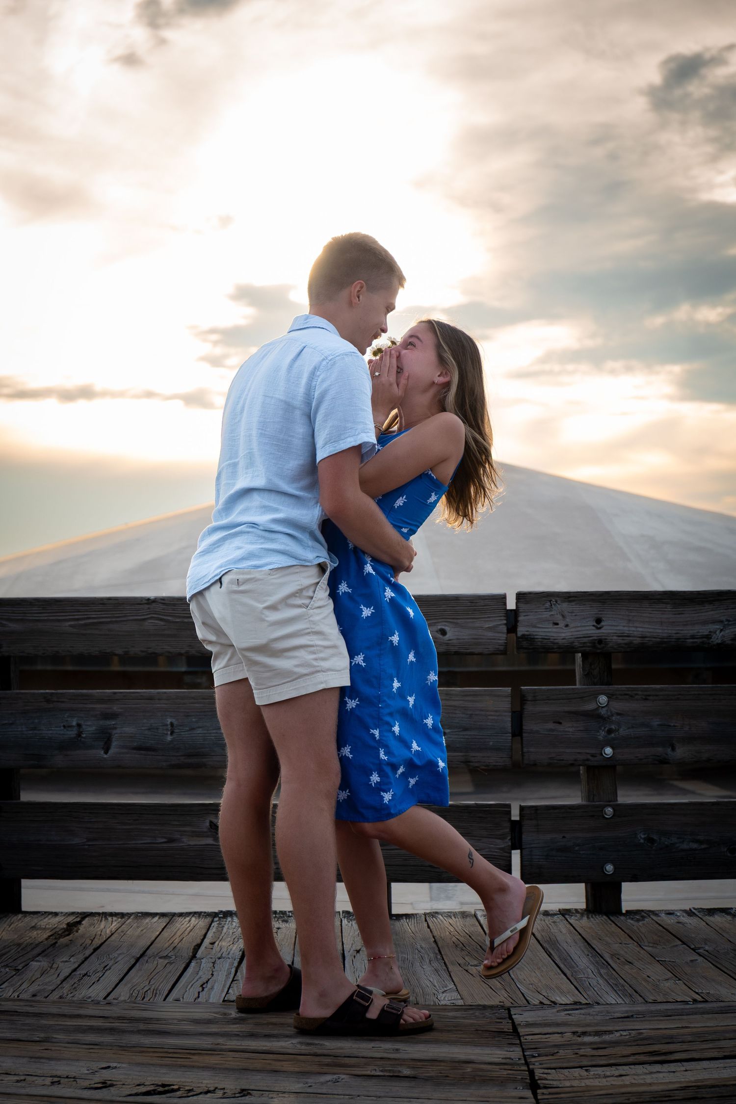 Couple embracing and almost kissing on a wooden pier at sunset, woman in blue dress, man in light blue shirt.