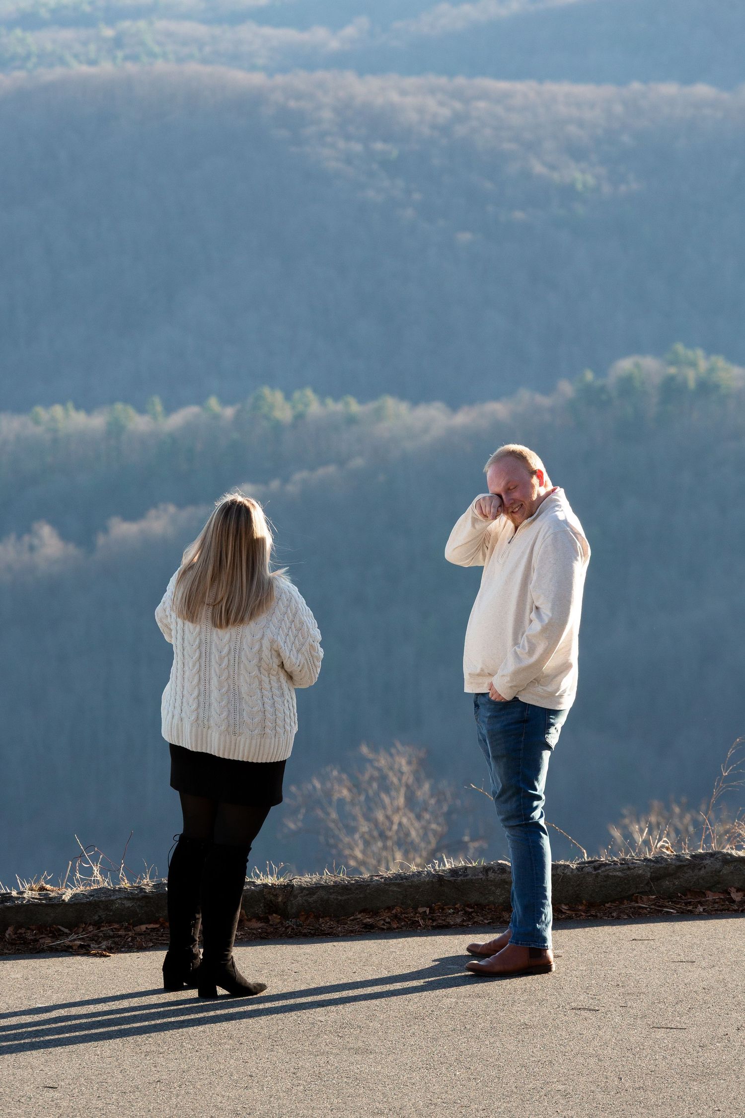 Two people in warm sweaters stand on a scenic mountain overlook on a sunny winter day with forested hills in background.