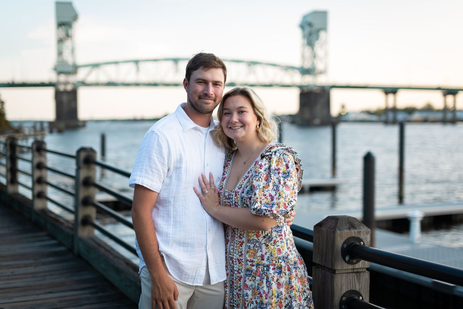 A smiling couple embraces on a wooden waterfront boardwalk with a vertical lift bridge and calm river in the background at sunset.