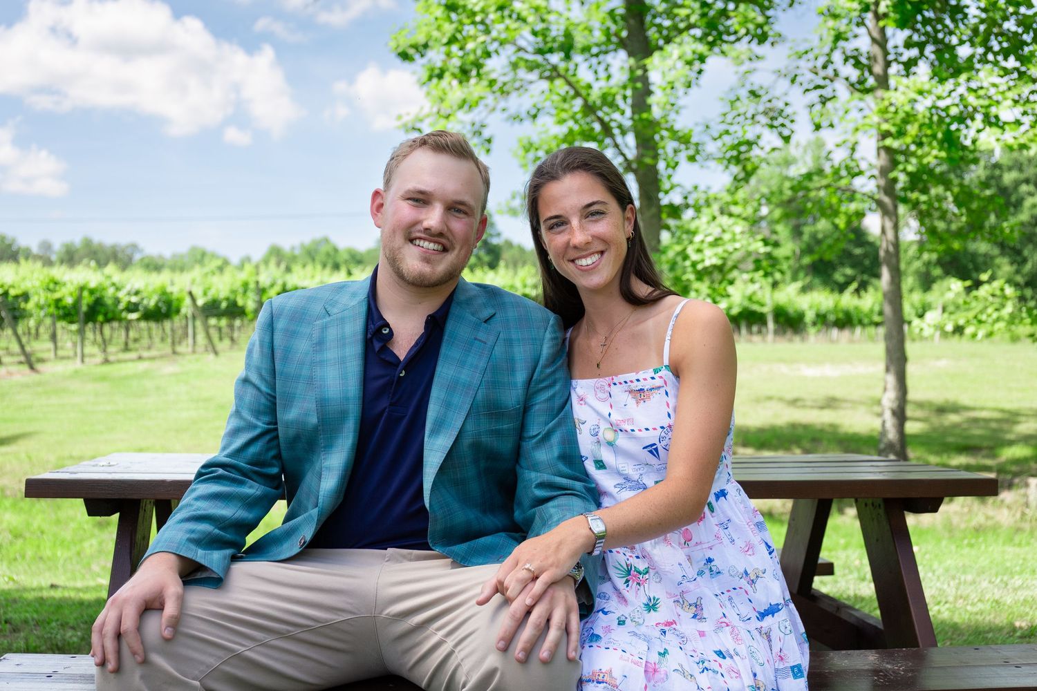 Smiling couple sitting at a picnic table at a vineyard on a sunny day, man in teal blazer, woman in floral dress.