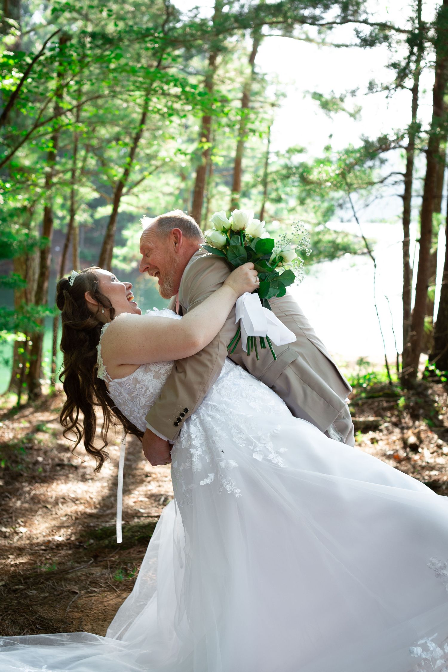 Bride in white gown and groom in tan suit share a joyful embrace in a sunlit forest near a lake.