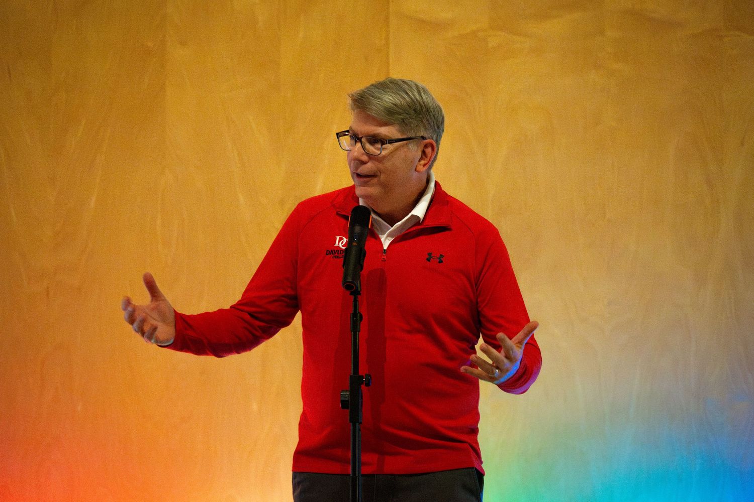 Speaker in a red sweater gestures while giving a presentation on stage.