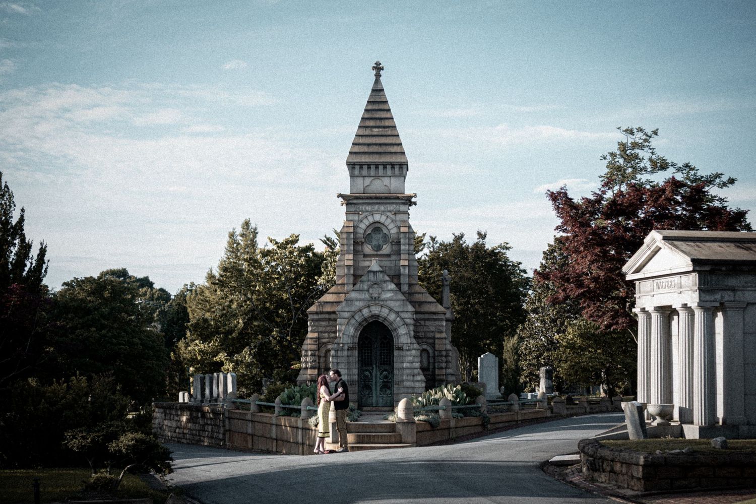 Couple standing before a grand Gothic stone mausoleum in a historic cemetery surrounded by lush trees.