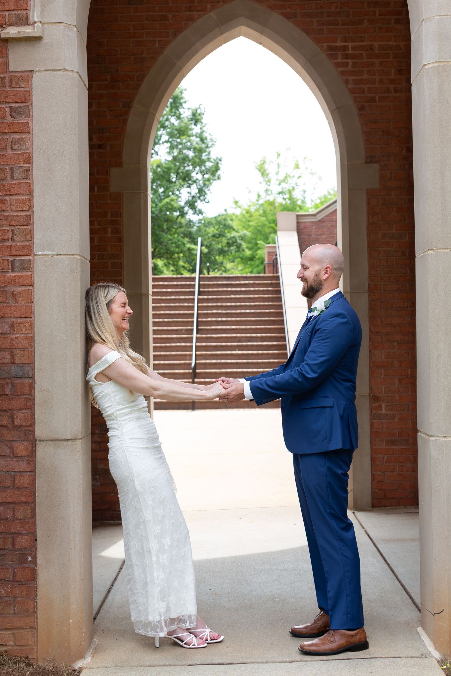 A bride and groom hold hands under a Gothic stone arch at a brick building, smiling at each other on their wedding day.