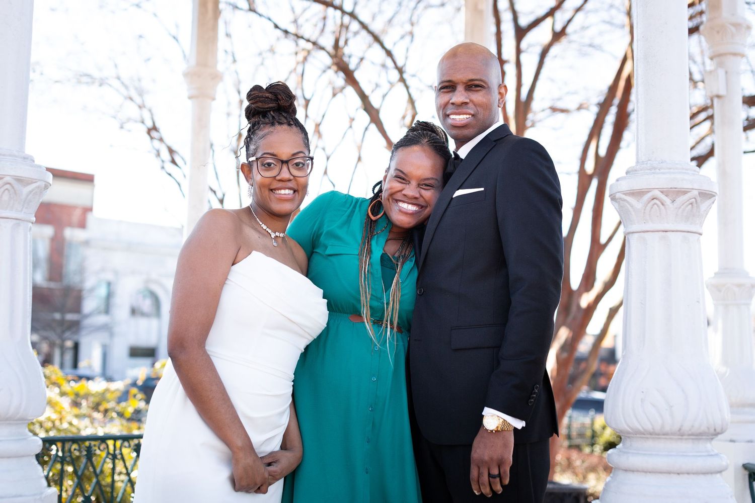 Wedding party members in formal attire pose together outdoors with white columns in background.