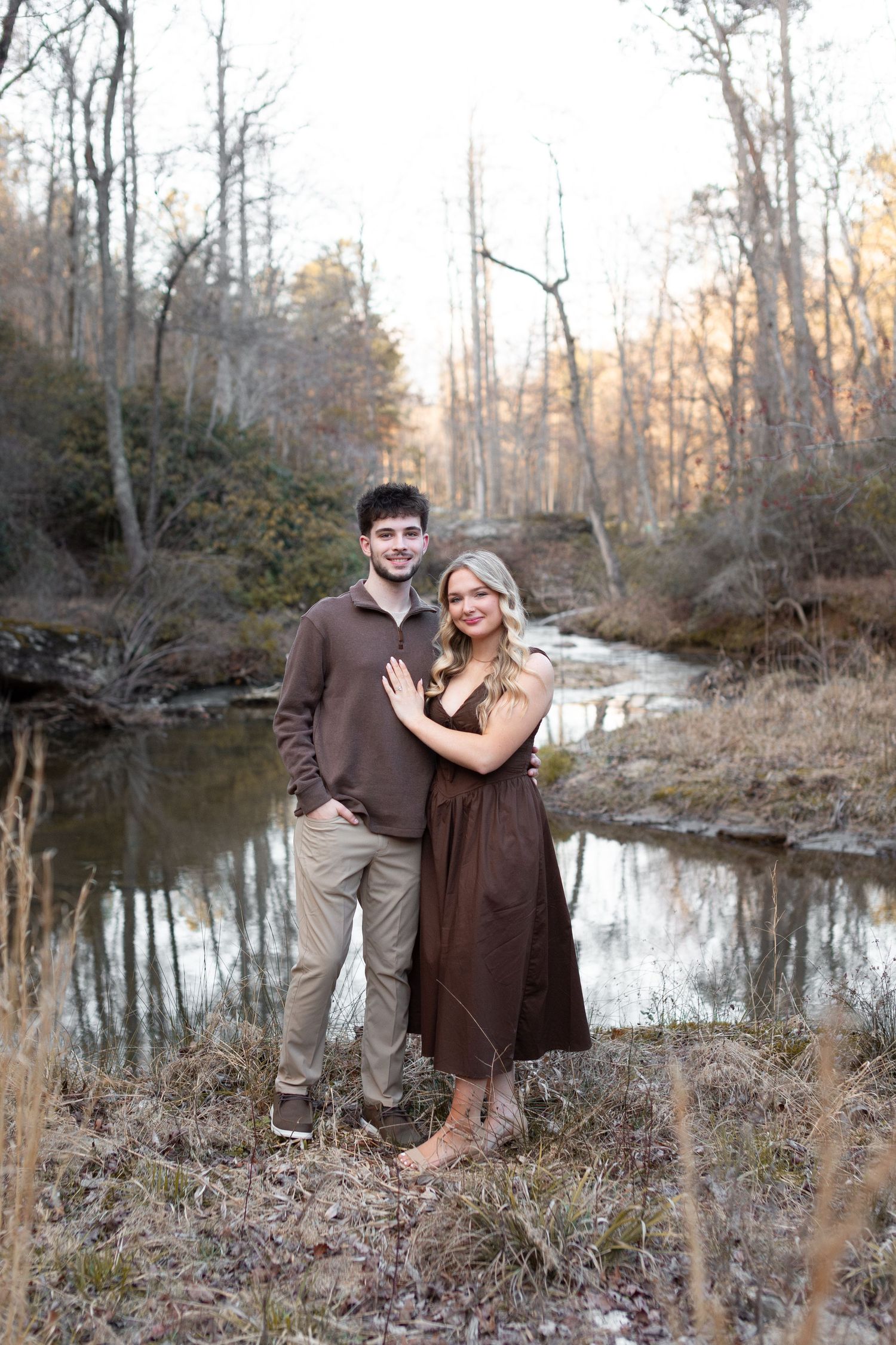 A couple in coordinating brown outfits poses together by a peaceful creek in an autumn forest setting.