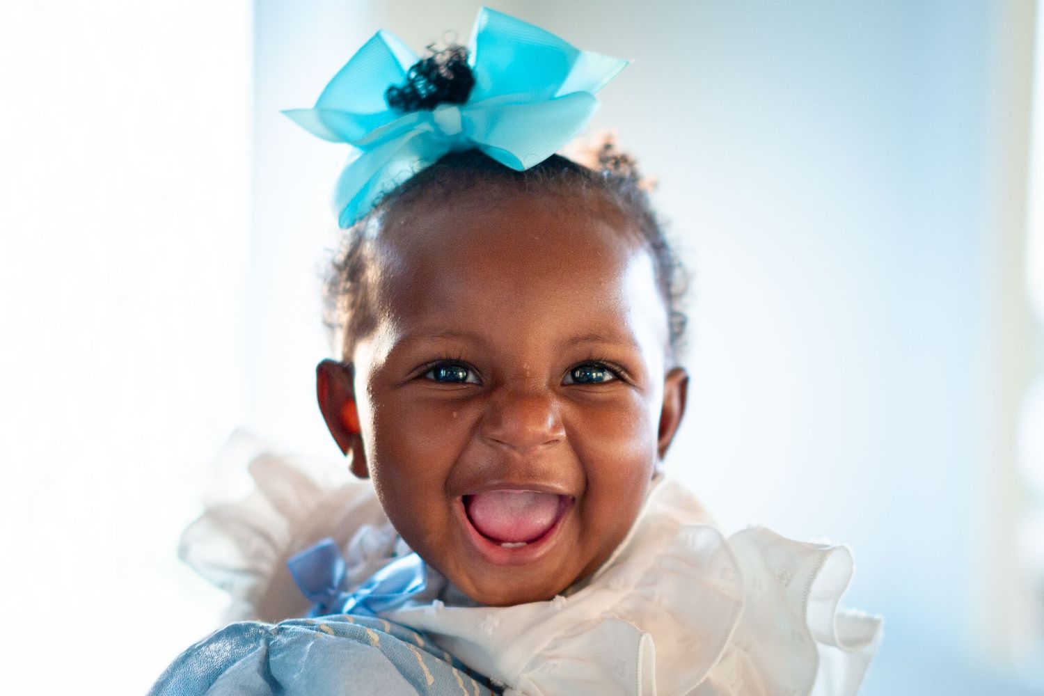 A happy baby in a white outfit and blue bow laughs with pure delight.