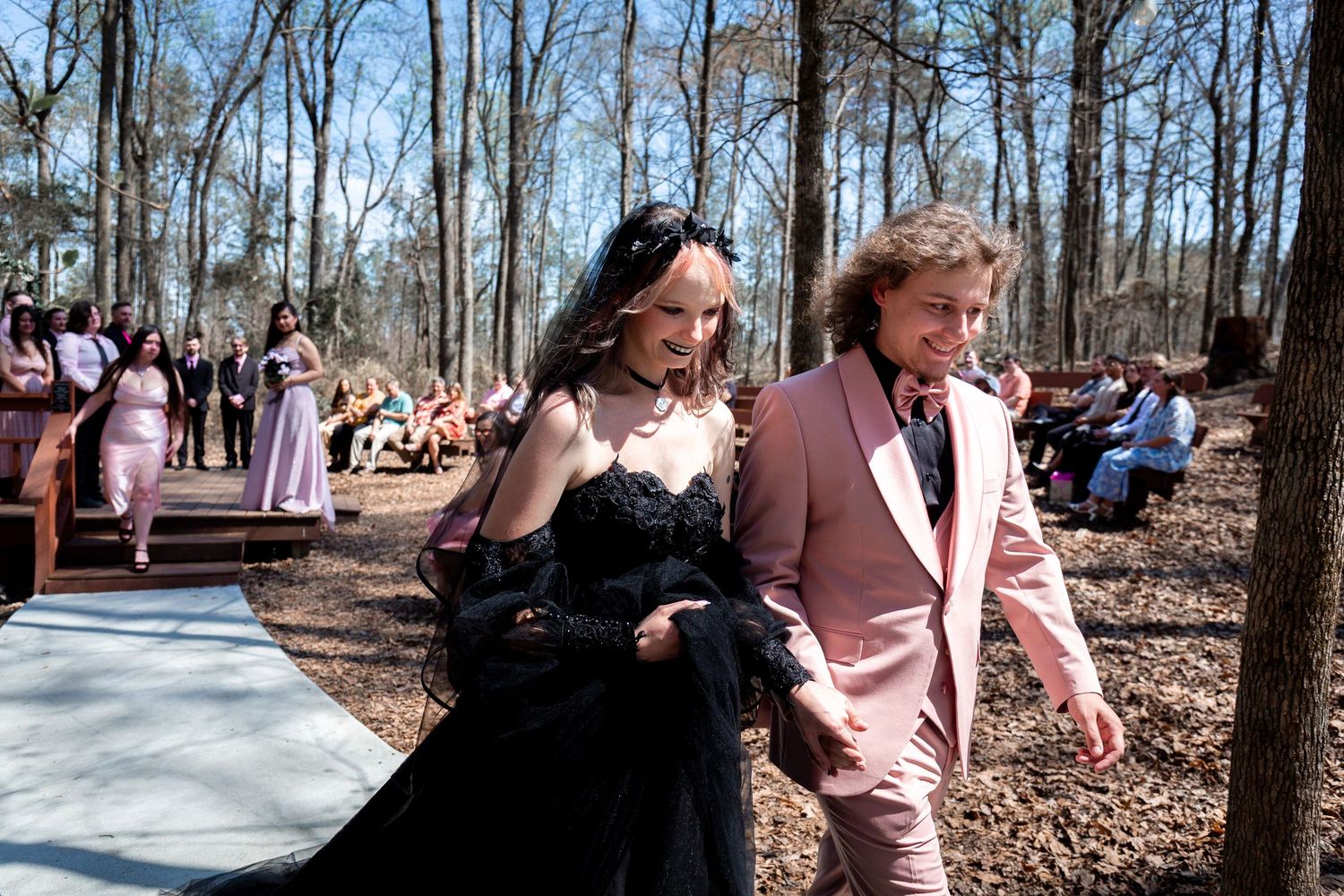 A group gathers outdoors in a wooded area on a sunny day wearing pink and casual attire for a social event.