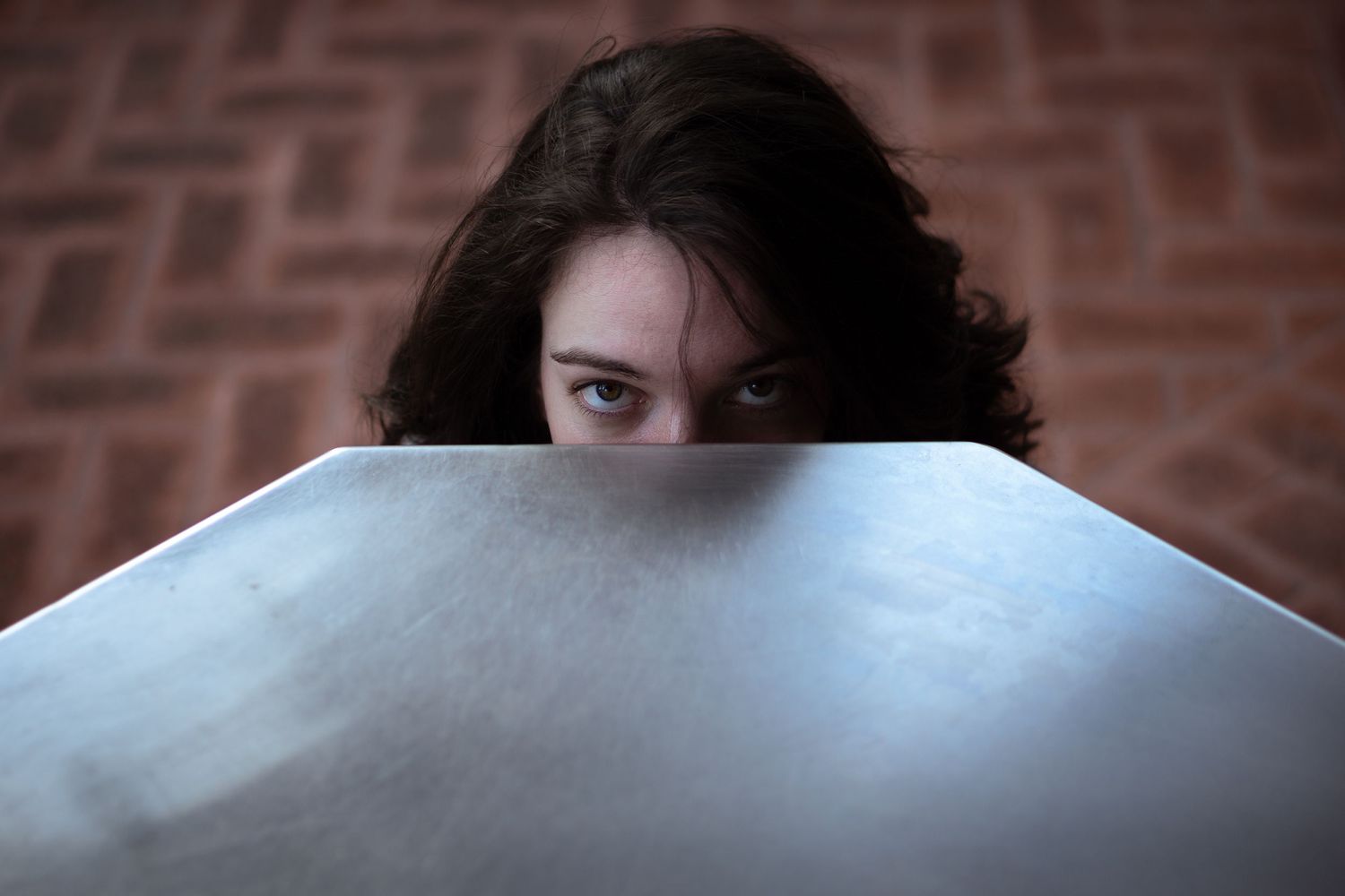 Young woman with curly dark hair peering over a laptop screen with curious green eyes against a brick wall.