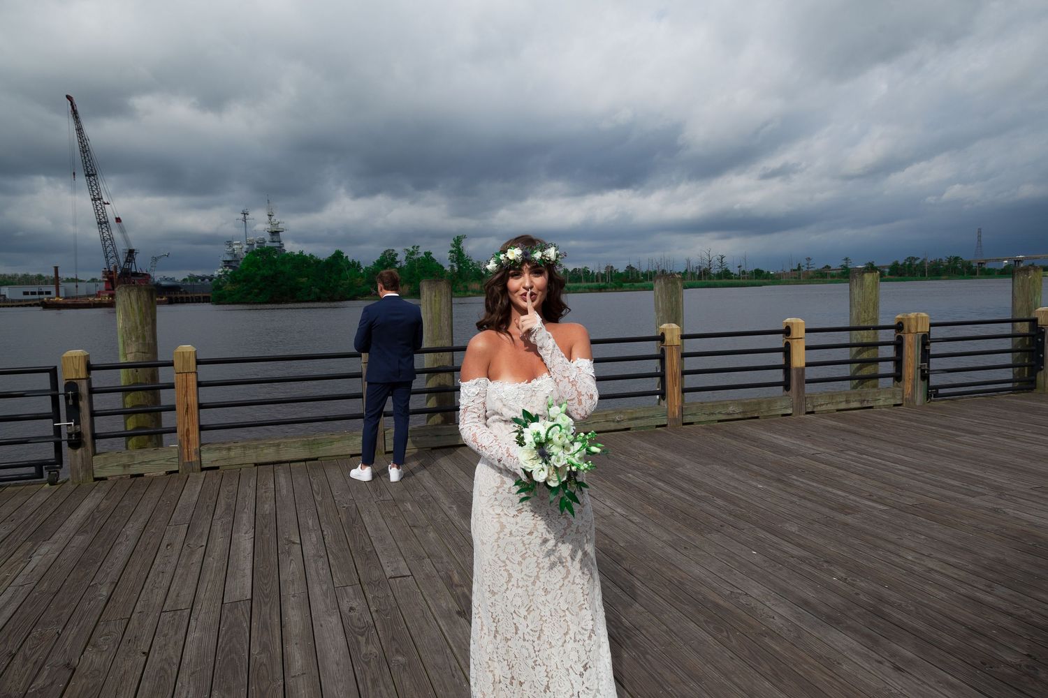 A bride in a white lace dress stands on a waterfront boardwalk under dramatic stormy clouds.