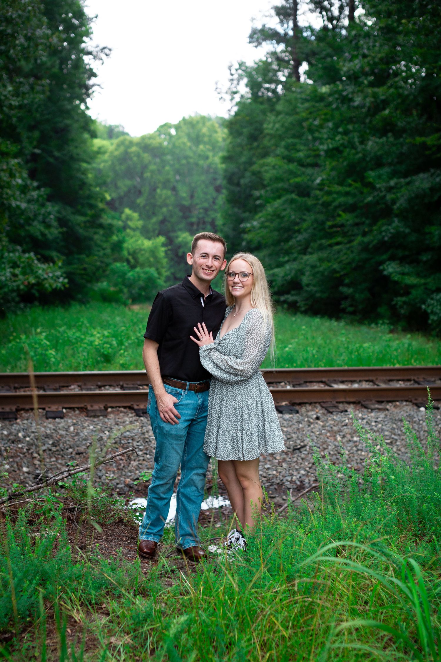 Engaged couple posing on railroad tracks surrounded by lush greenery.
