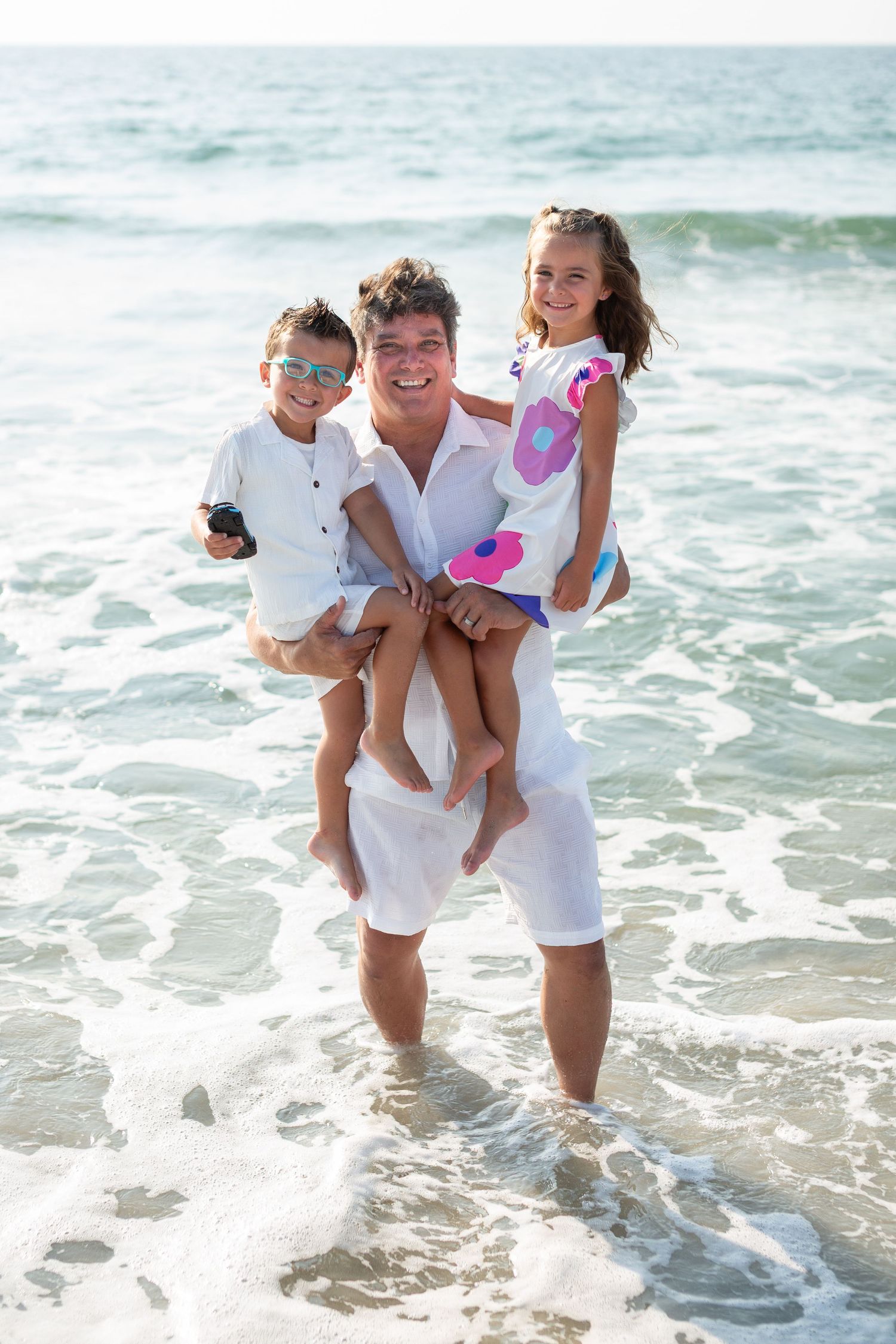 Parent holding two children while standing in ocean waves wearing white summer clothing.