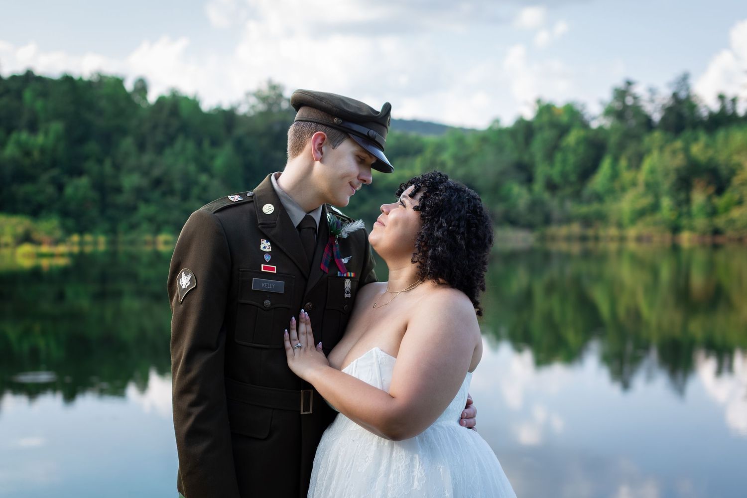 A smiling groom in a military dress uniform and a bride in a white gown embrace beside a calm lake with green trees reflected in the water.