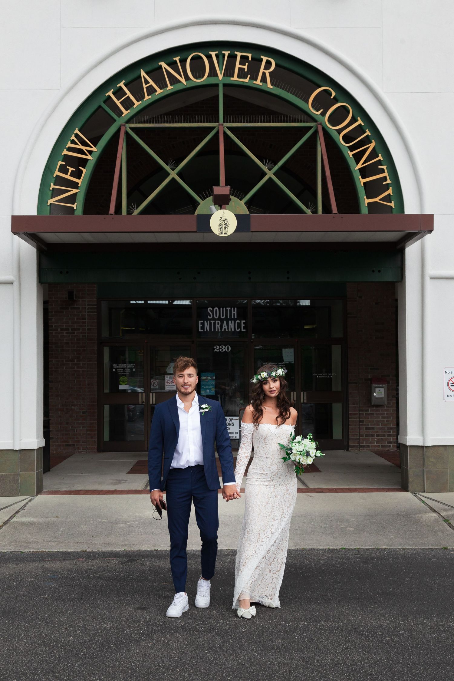 A couple walks together through an elegant arched entrance with white columns and dark green signage.