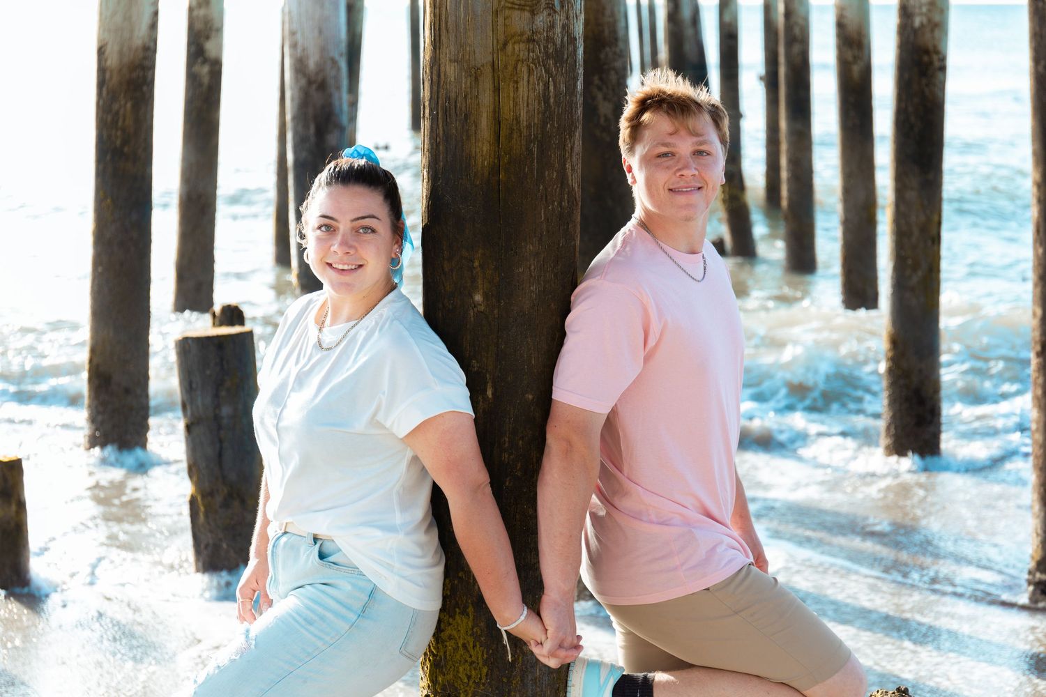 A couple shares a tender moment on a sunny beach boardwalk with wooden pier posts in the background.