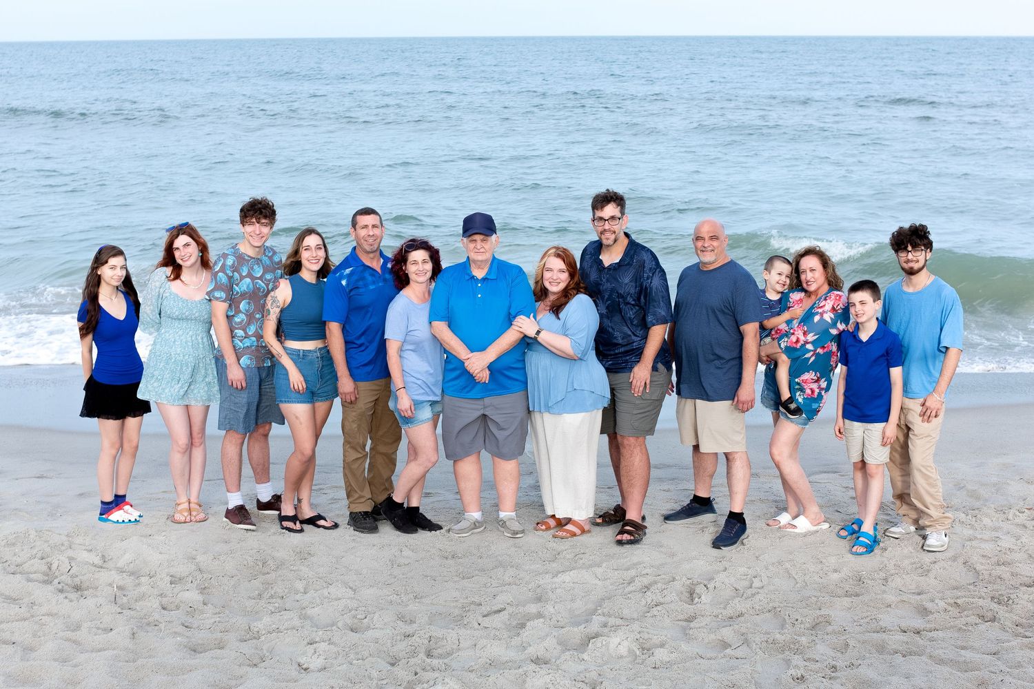 Large family group portrait on sandy beach wearing coordinated blue and white outfits.