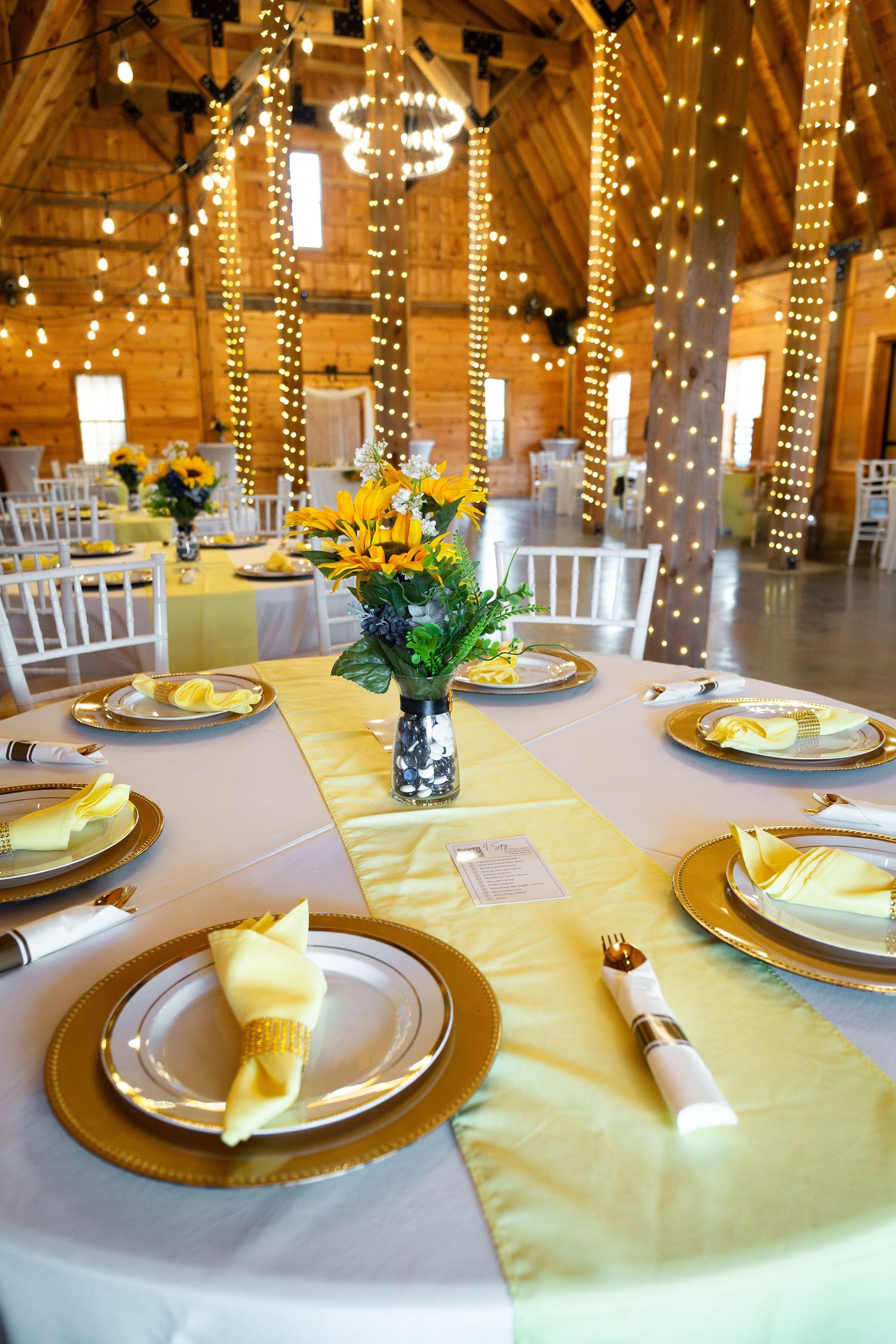 Elegant wedding reception table with yellow linens, gold chargers and hanging string lights in a rustic barn.