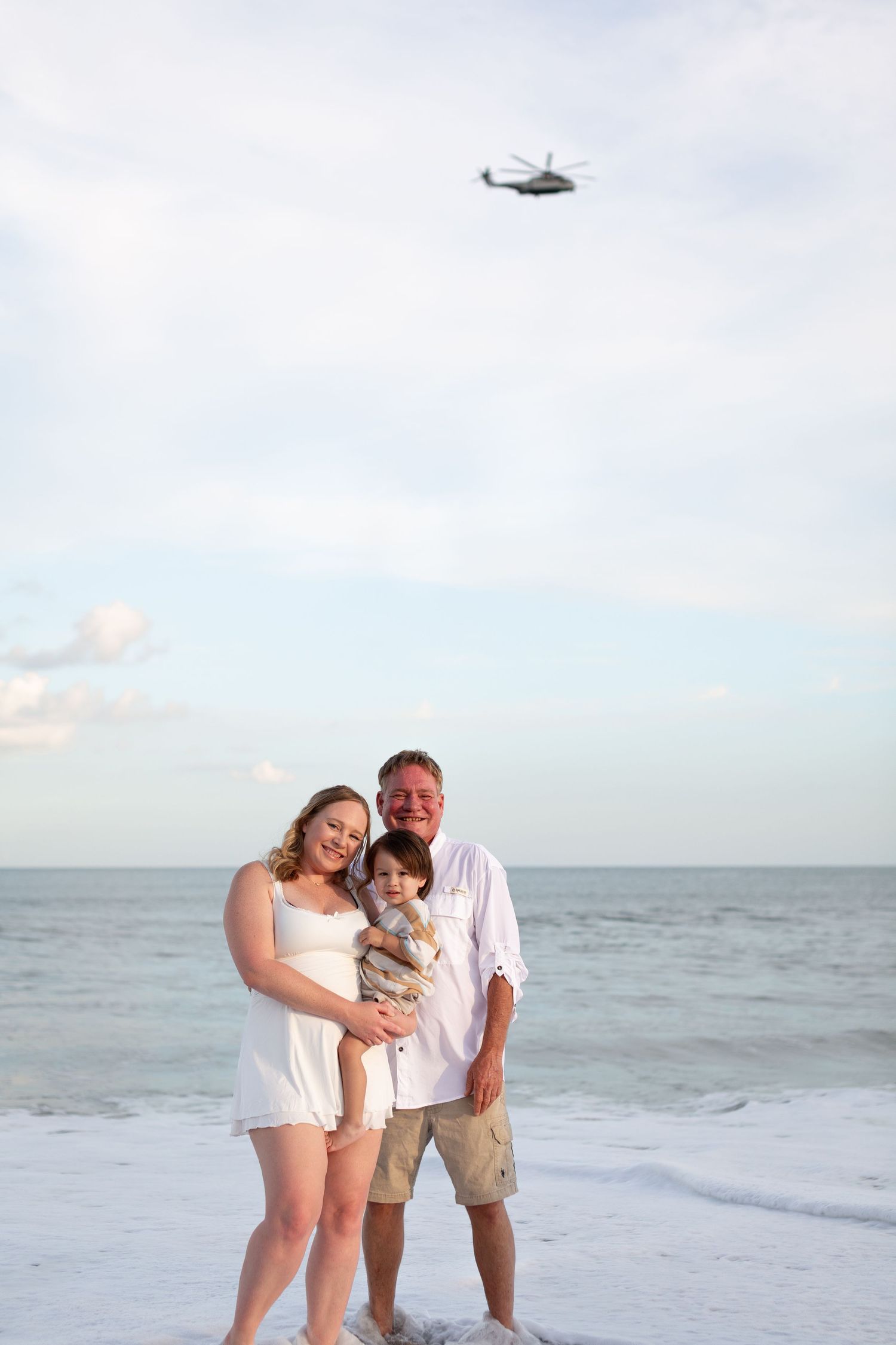 Family of three poses on a beach with ocean waves, a helicopter flying overhead in the cloudy sky.