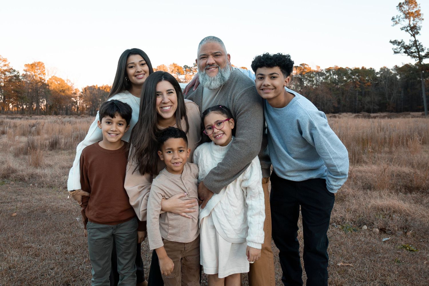 A happy family of seven poses together in a sunlit field with trees in the background during golden hour.