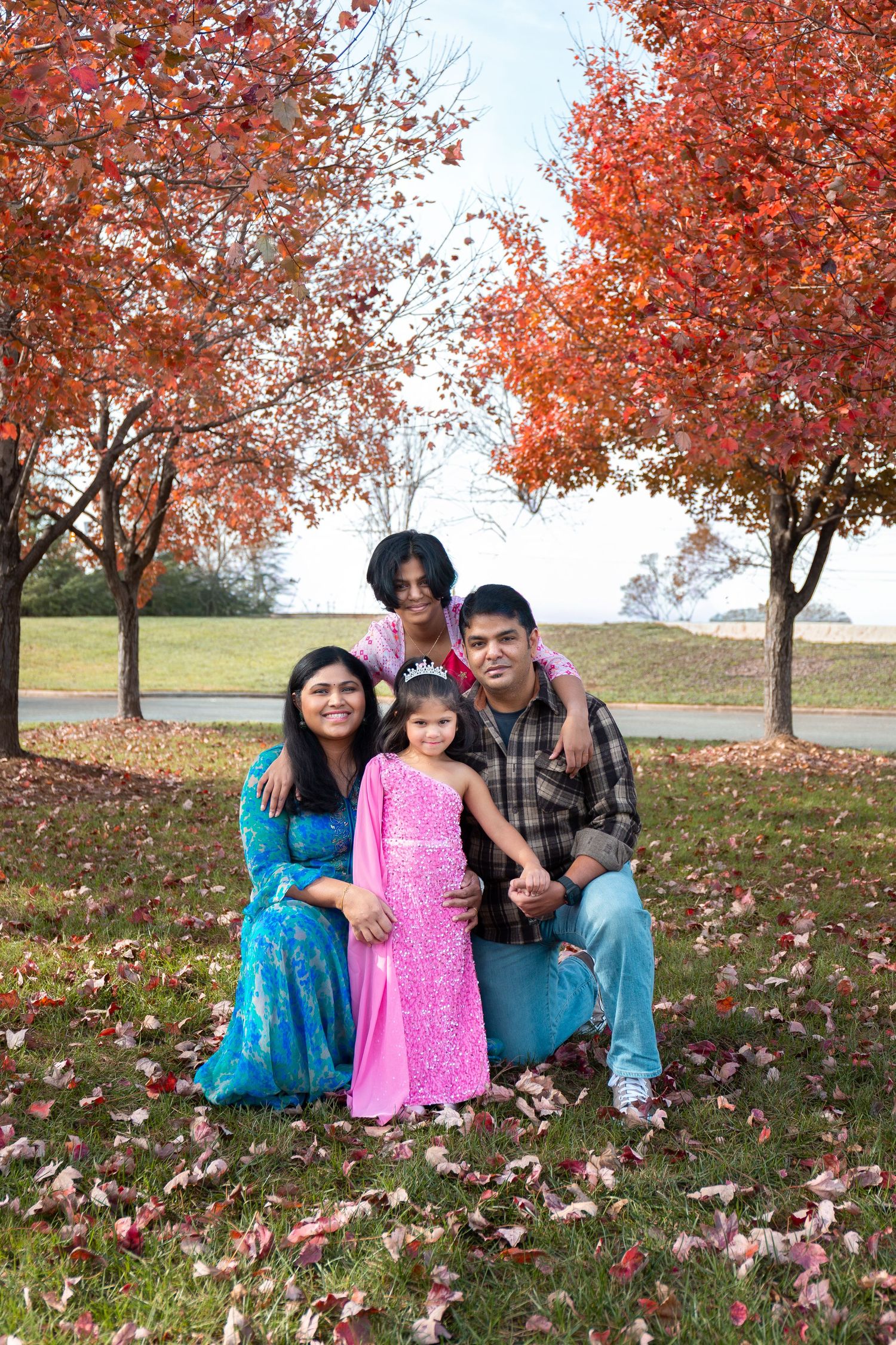Family sitting together under vibrant red autumn trees with fallen leaves scattered on the ground.
