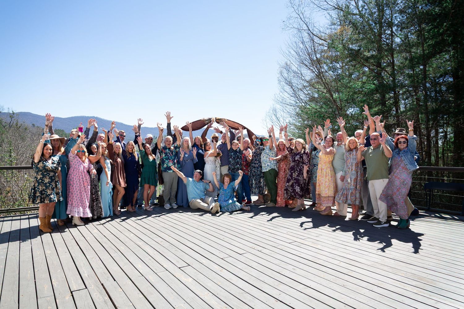 Large group of people posing together on an outdoor wooden deck with mountains visible in the background.