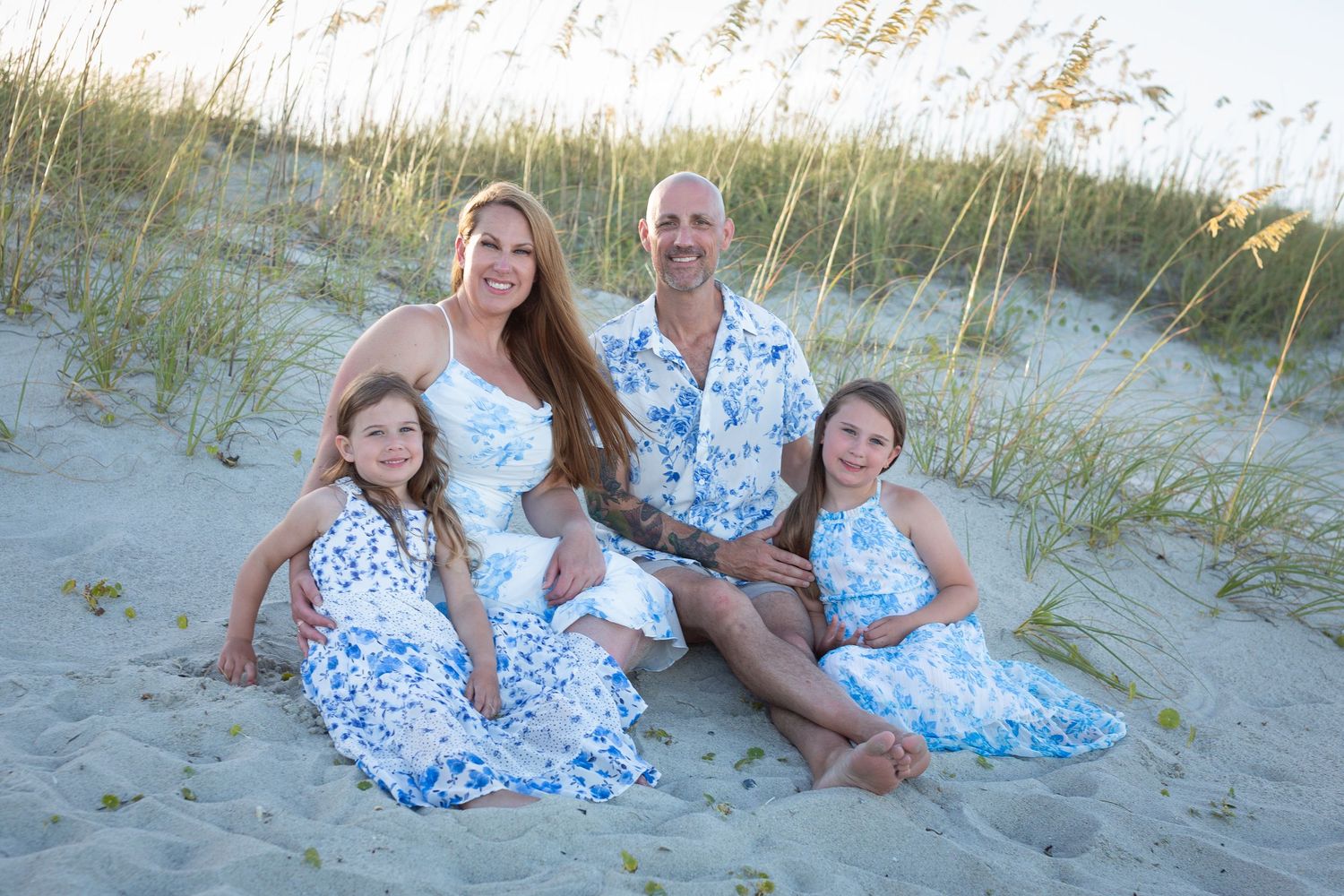 A family group wearing coordinated blue and white outfits poses together on a sandy beach with sea grass in the background.