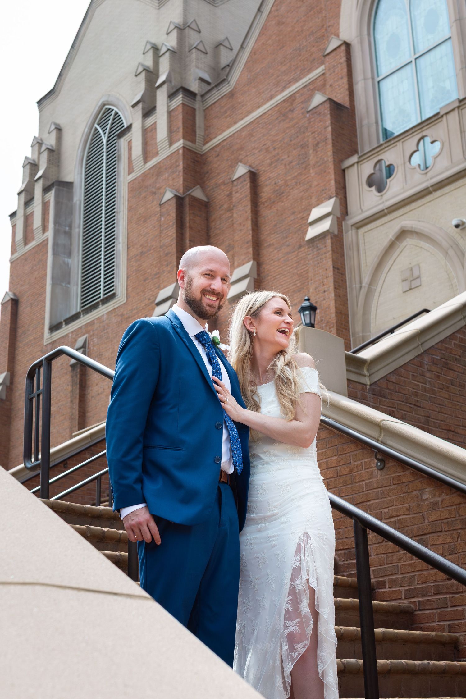 Bride and groom laughing together on church steps, groom in blue suit, bride in white lace dress.