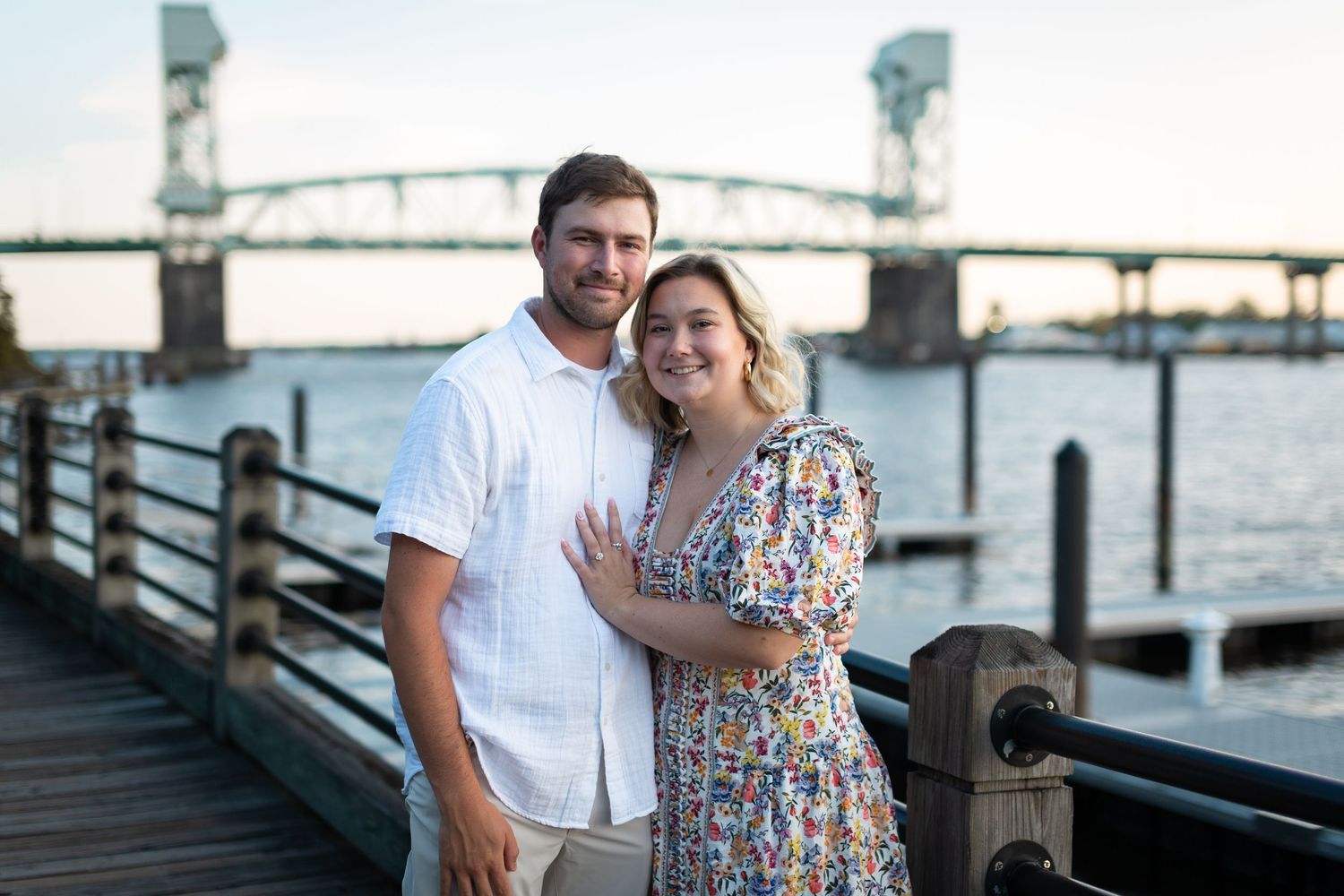 A couple poses on a waterfront boardwalk with a bridge and industrial cranes visible in the background.