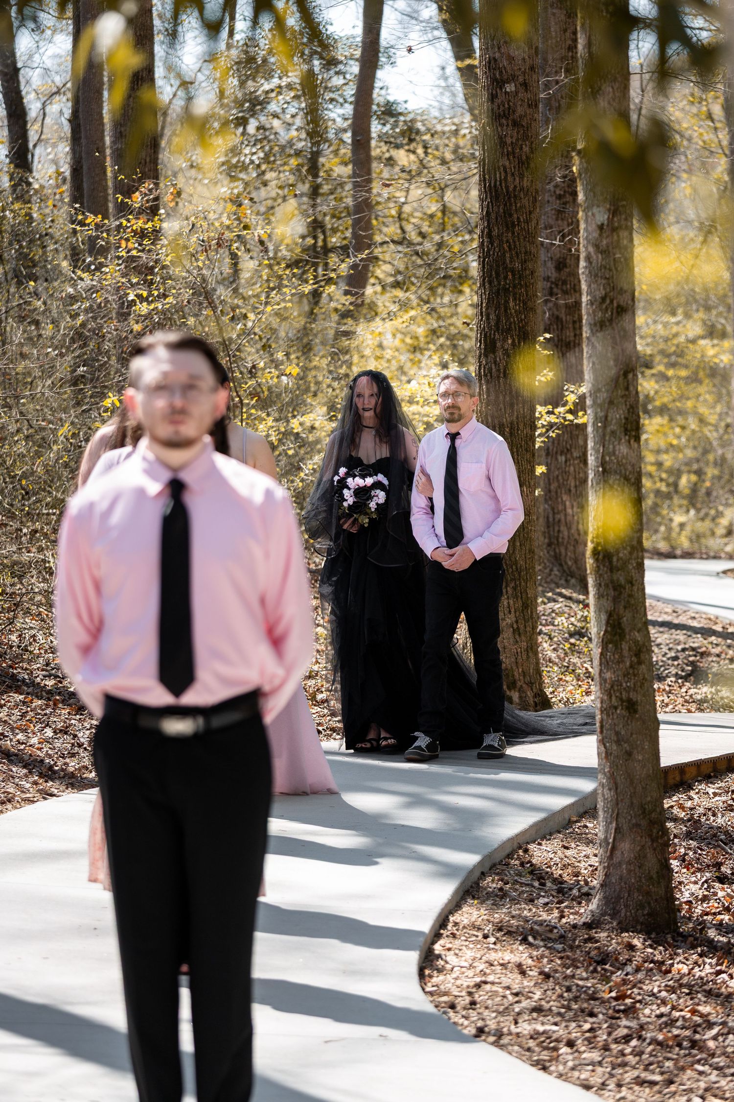 A group of people in pink shirts and black ties pose for photos in a wooded outdoor setting with autumn foliage.